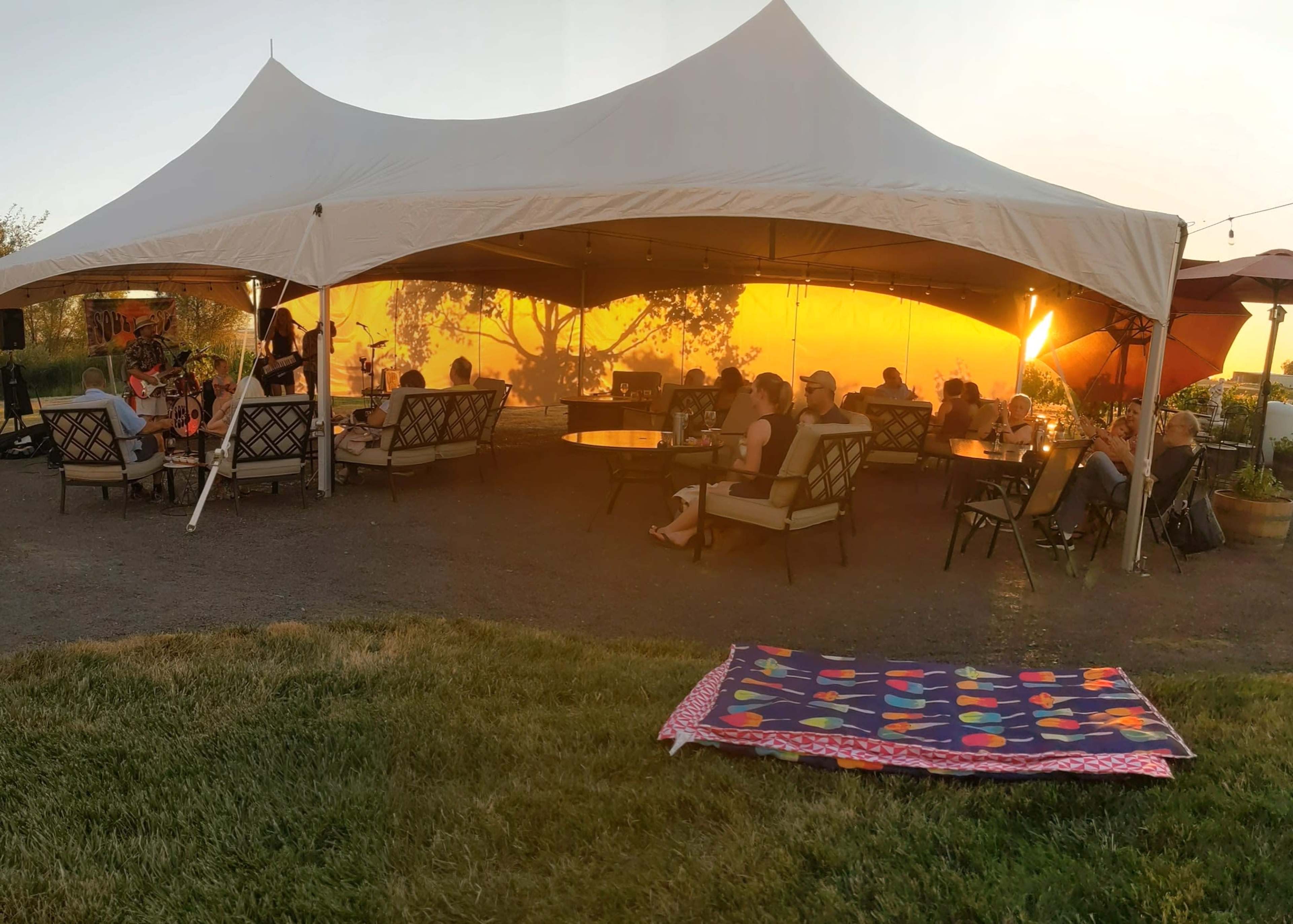 A large tent hosts an outdoor gathering with people seated on sofas, while a colorful quilt lies on the grass nearby.