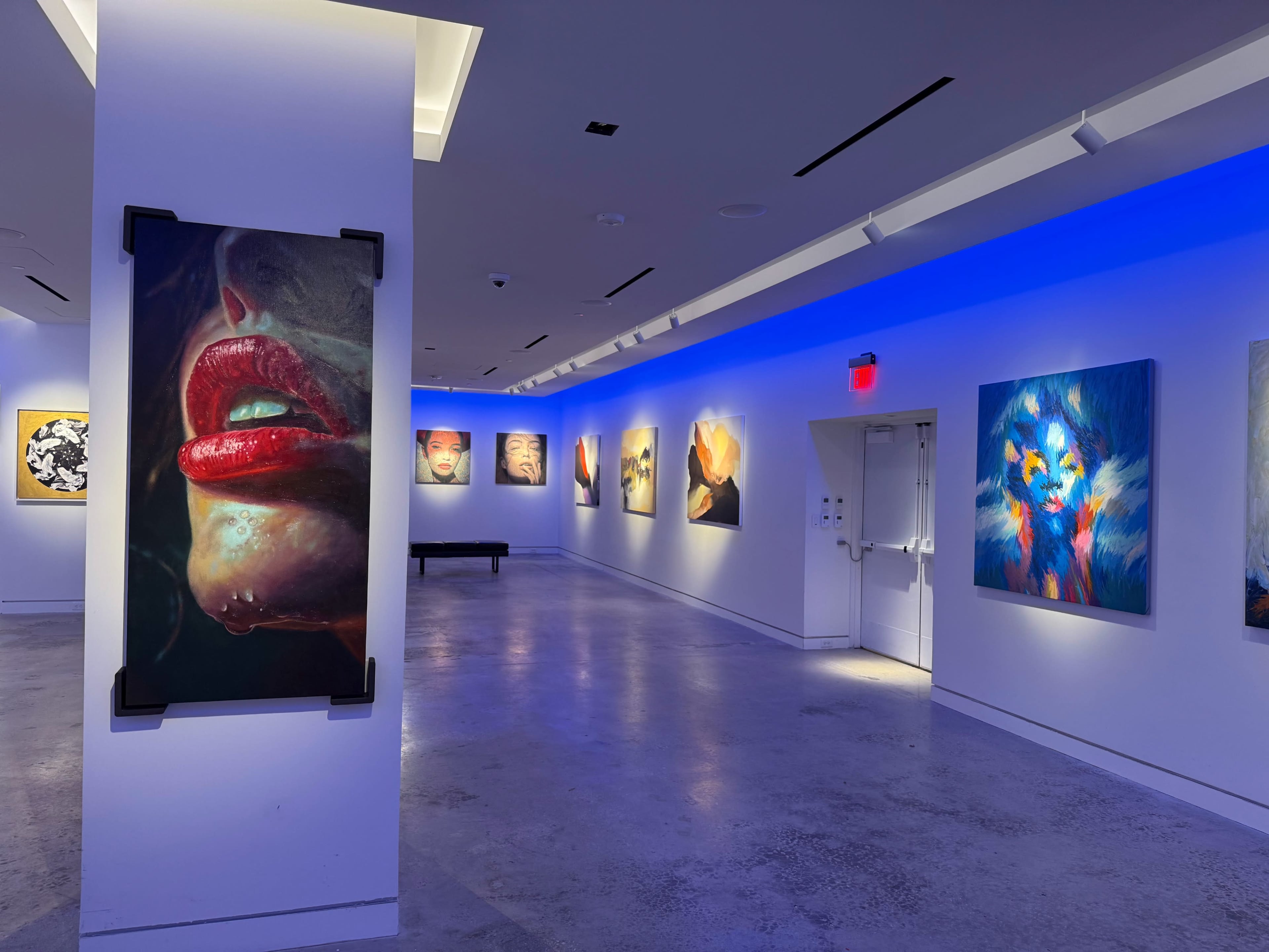 The image shows an art gallery with colorful paintings displayed on white walls, illuminated by blue lighting.