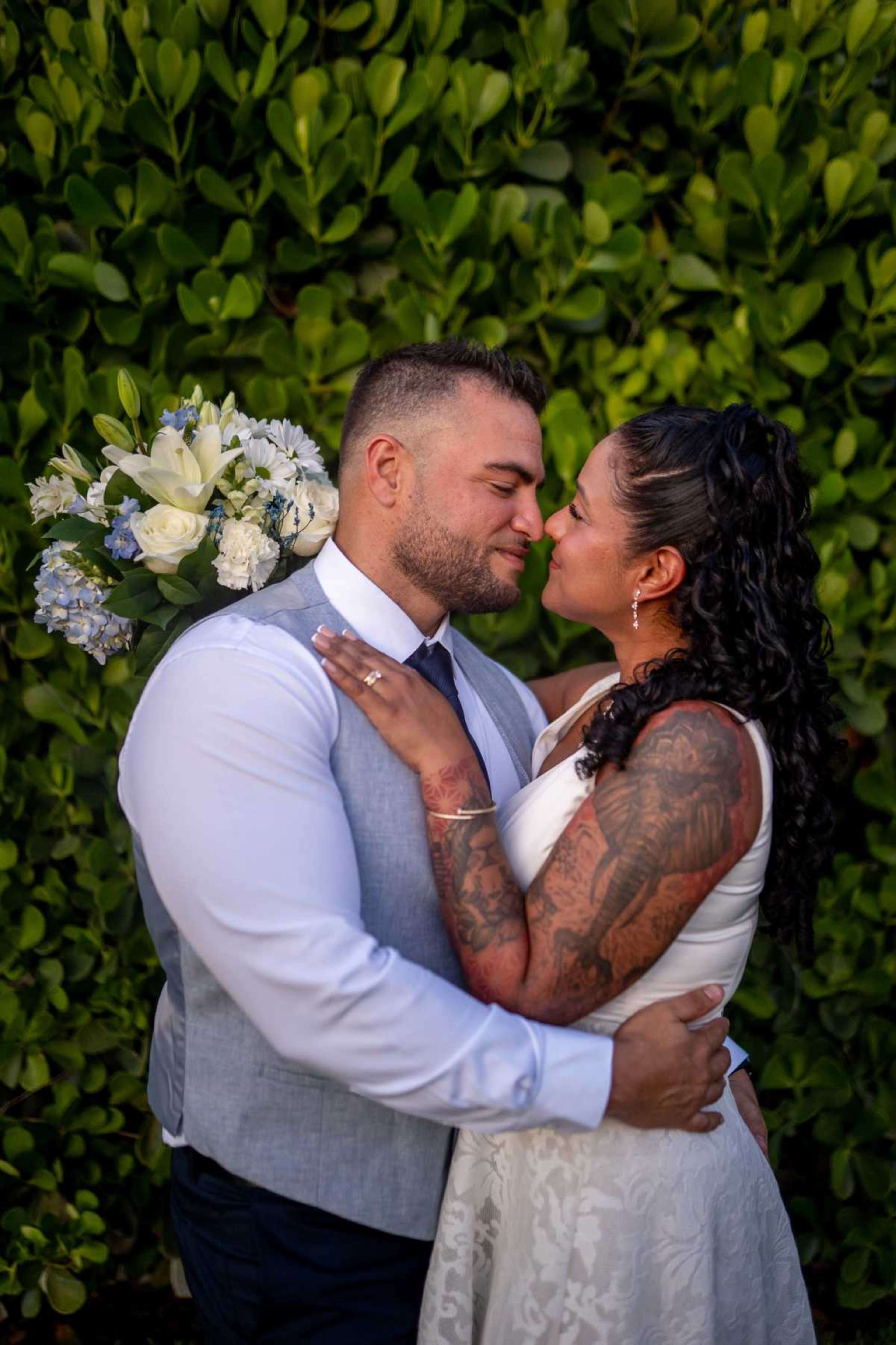 A couple embraces and kisses, surrounded by a lush green backdrop, while holding a bouquet of flowers.