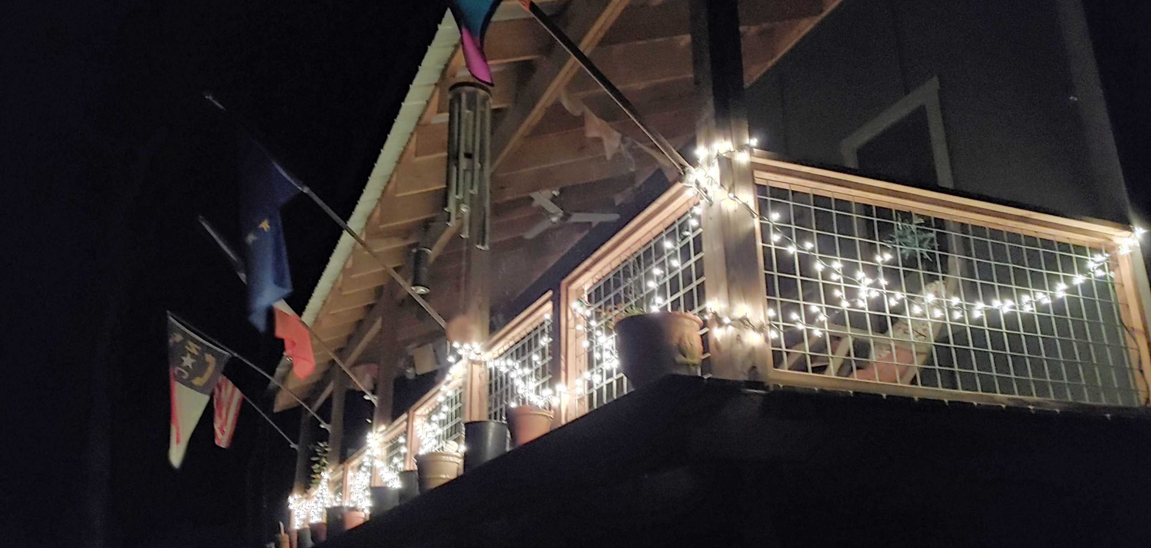 A wooden balcony illuminated by string lights features several potted plants and flags hanging from the rafters.