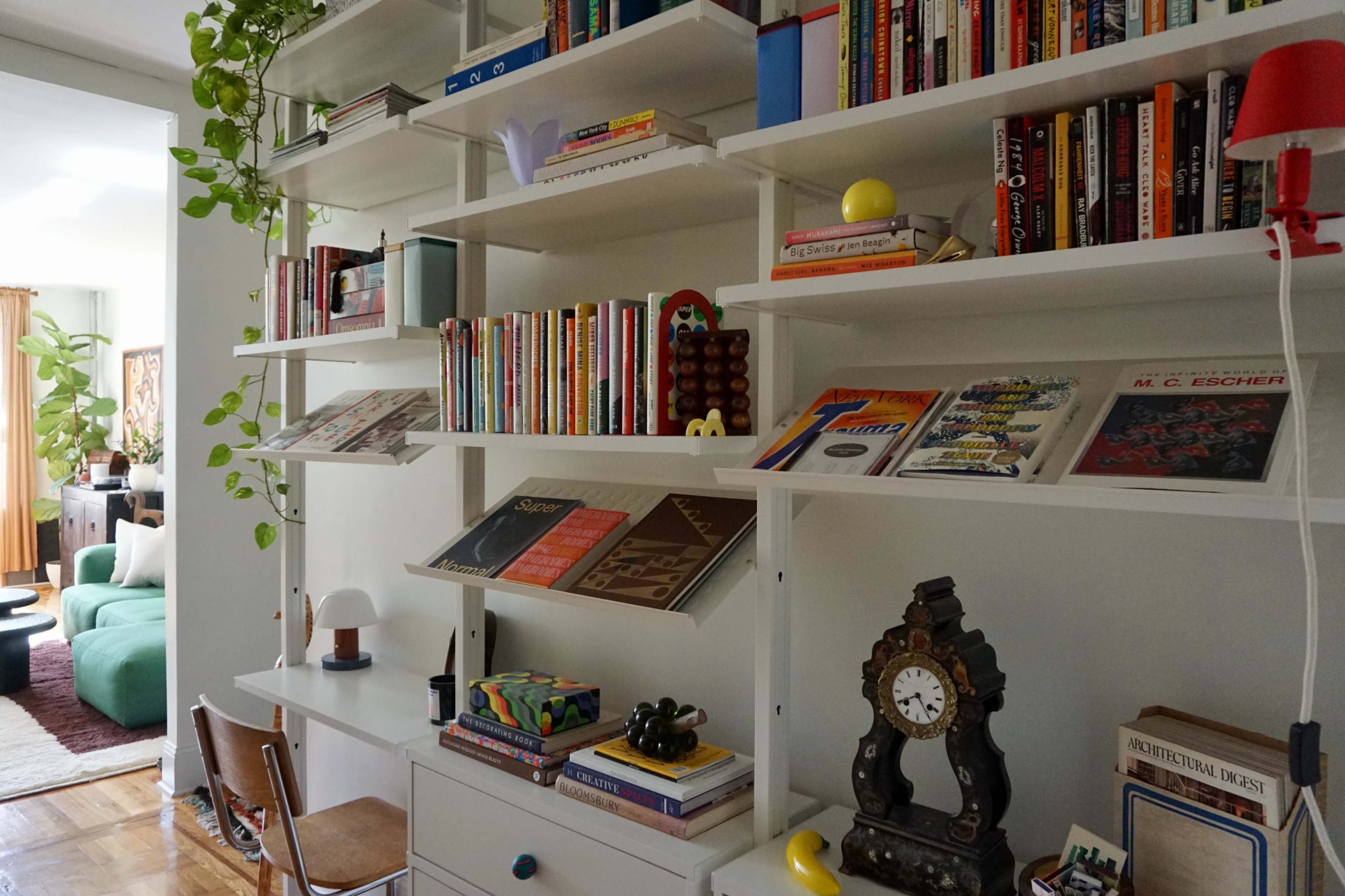A modern bookshelf filled with an array of books, decorative items, and a clock, set against a bright interior with a glimpse of a living area in the background.