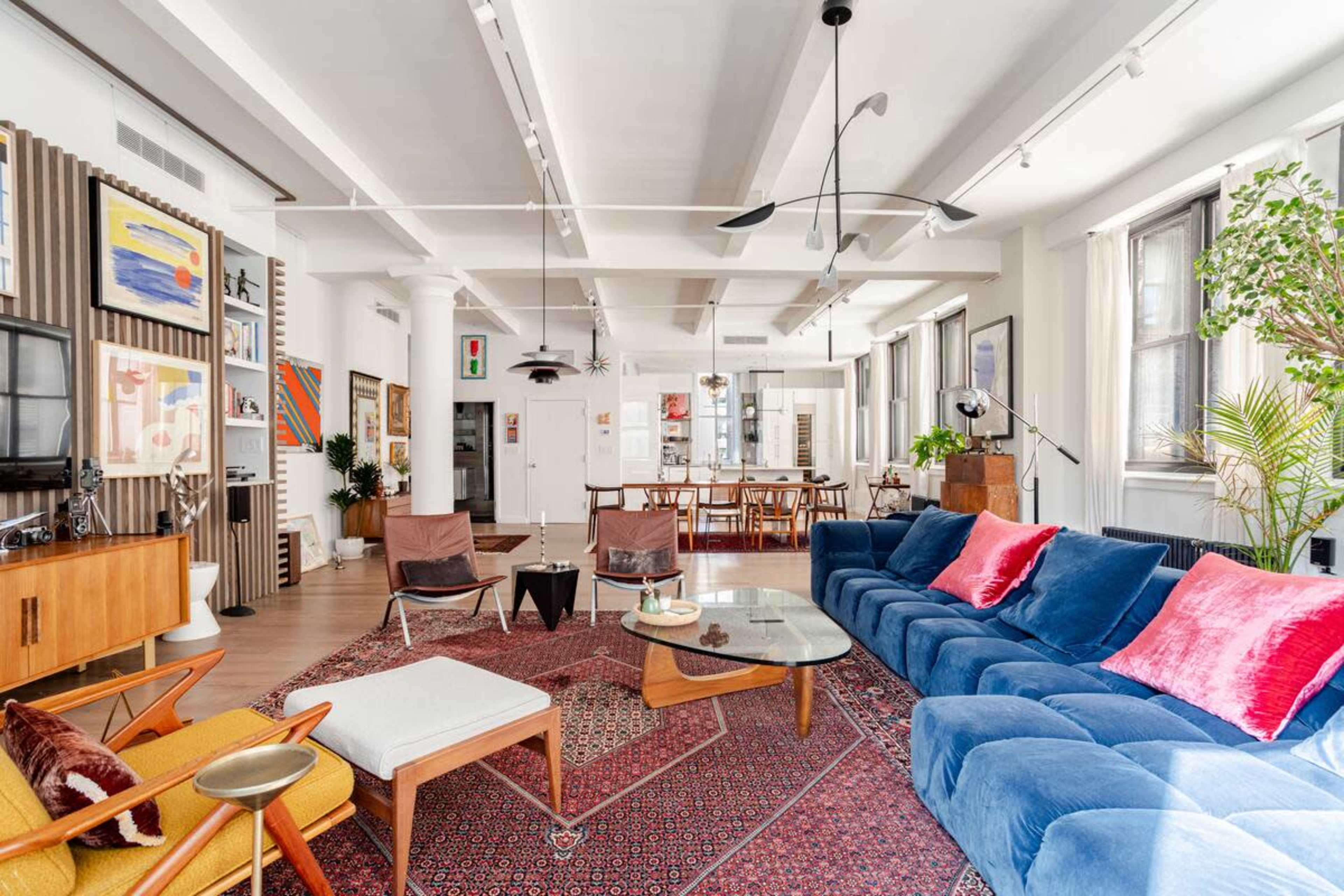 The image shows a spacious living room with a blue sectional sofa, wooden furniture, a large area rug, and various decorative artwork on the walls.