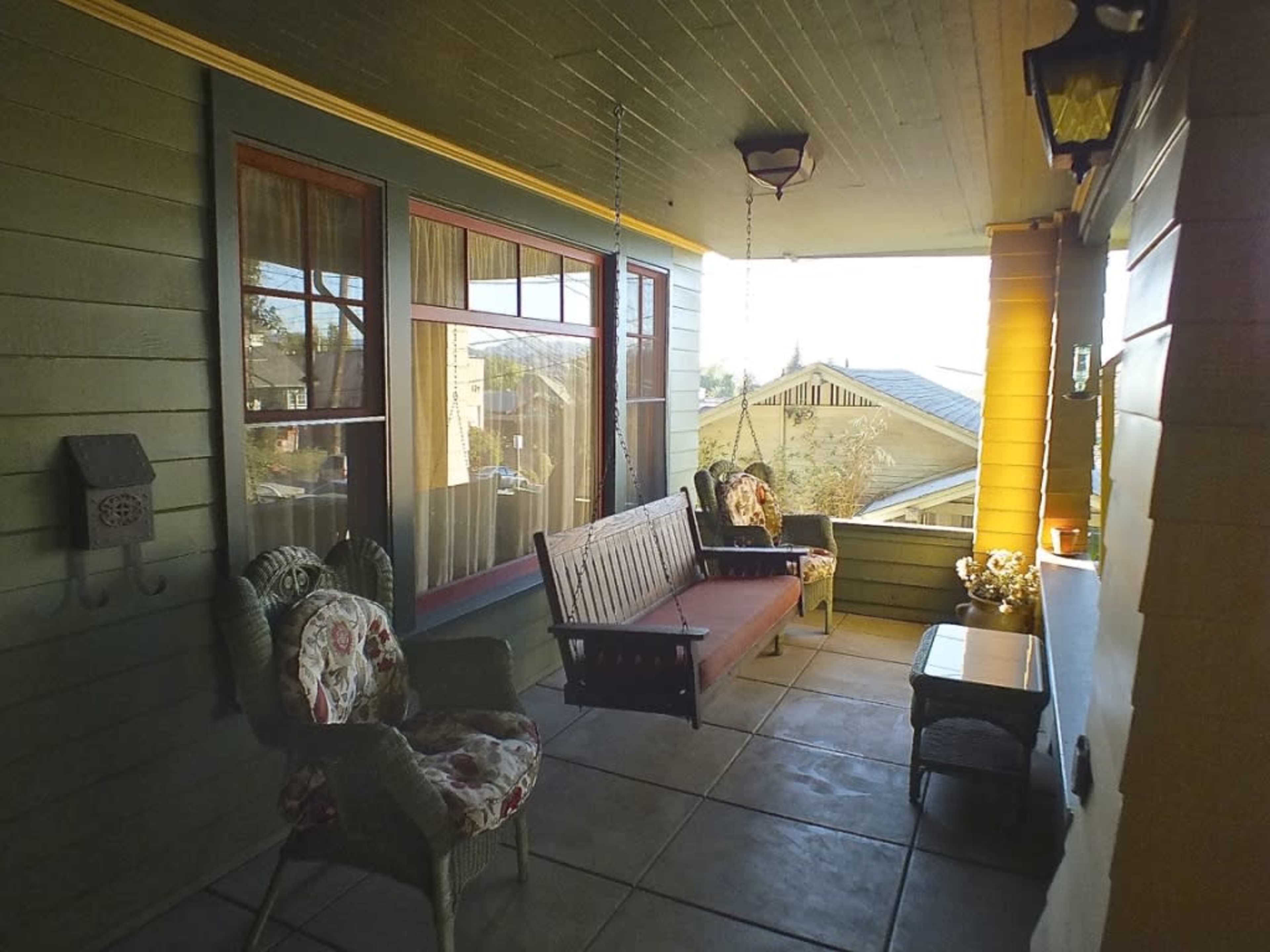 A cozy porch with a swing bench, a floral-patterned chair, and a small table, all set against a backdrop of green walls and large windows.