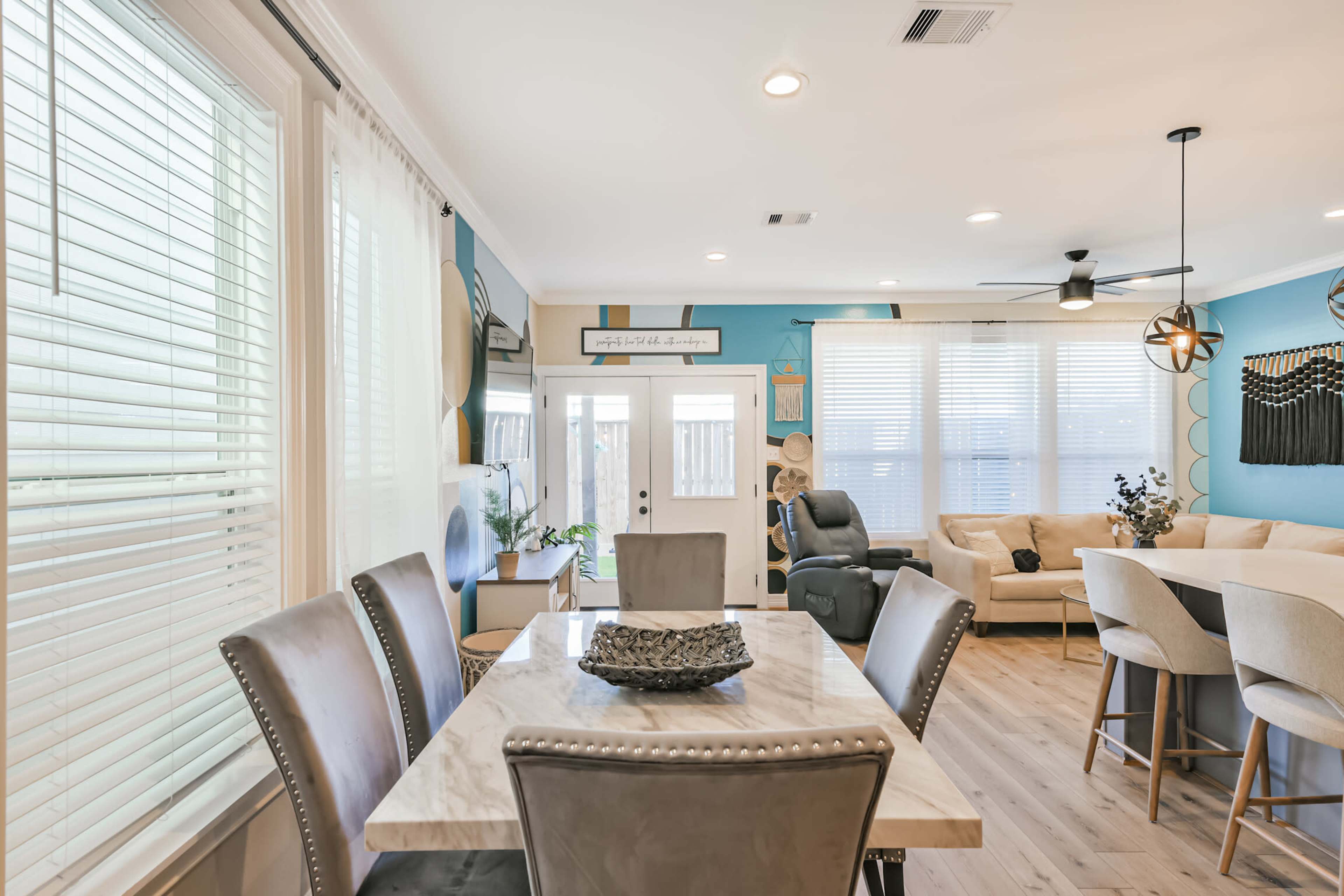The image shows a dining area with a marble table and upholstered chairs, leading into a brightly lit living space featuring a sofa, ceiling fan, and large windows.
