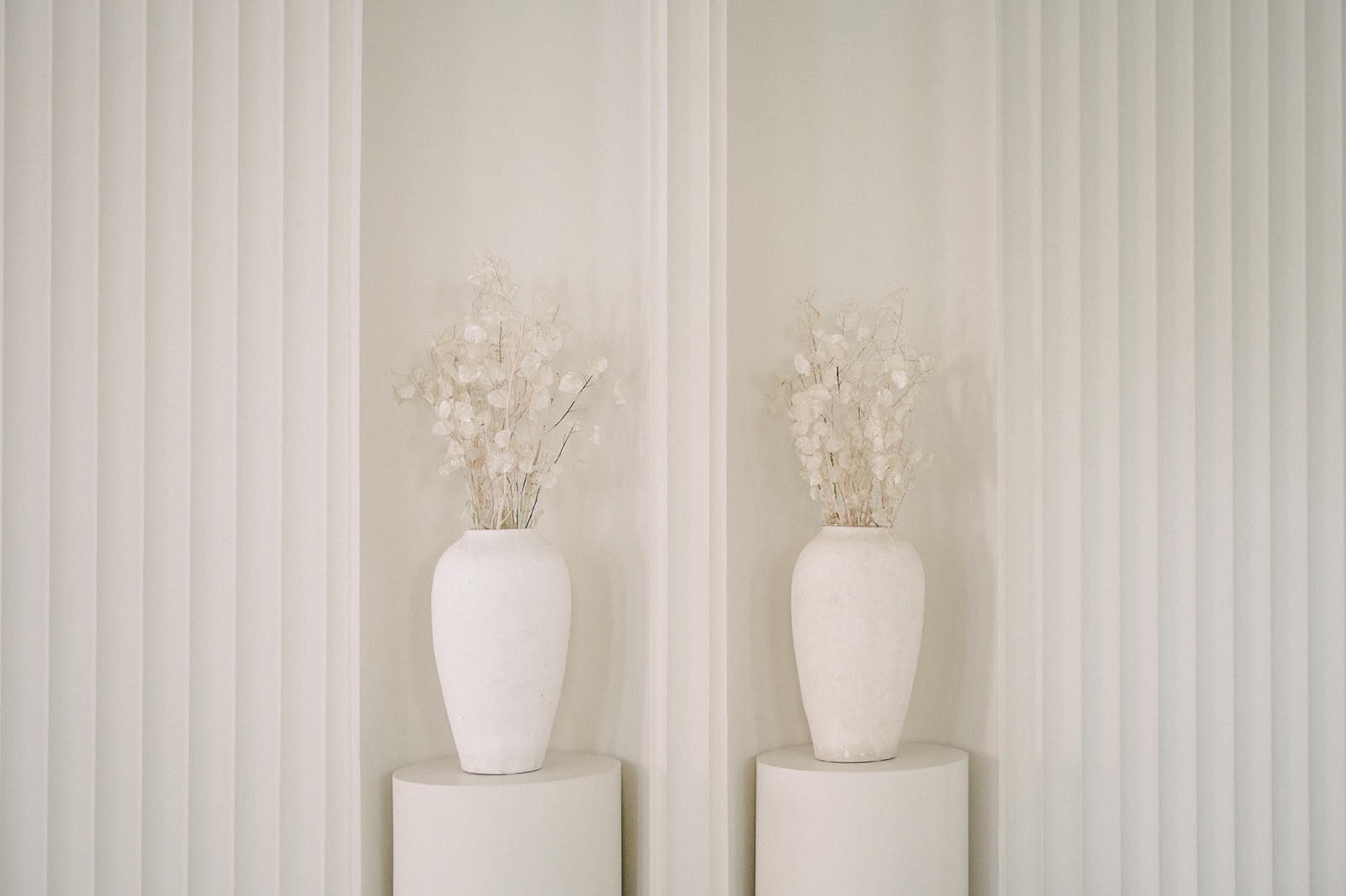 Two white vases filled with dried flowers are placed on cylindrical pedestals against a backdrop of vertical white panels.