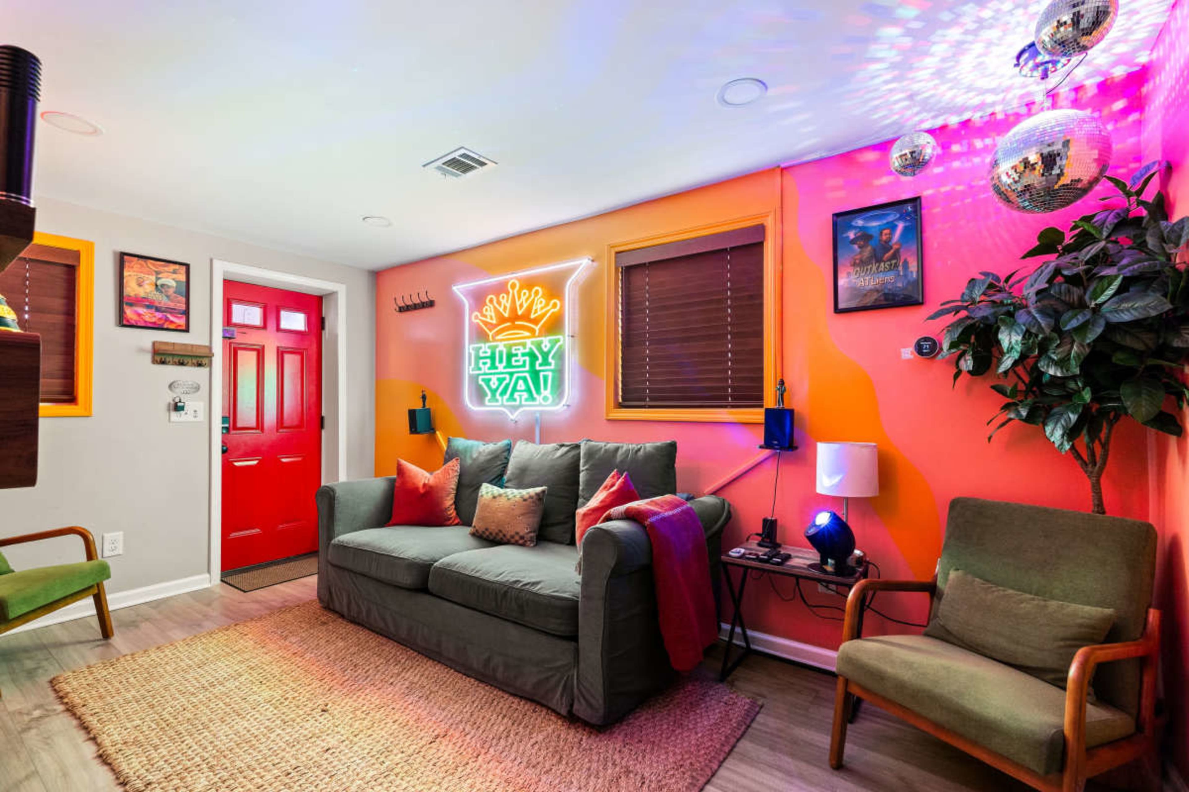 The image shows a brightly colored living room featuring a red front door, a green couch with decorative pillows, and a neon sign on a wall.