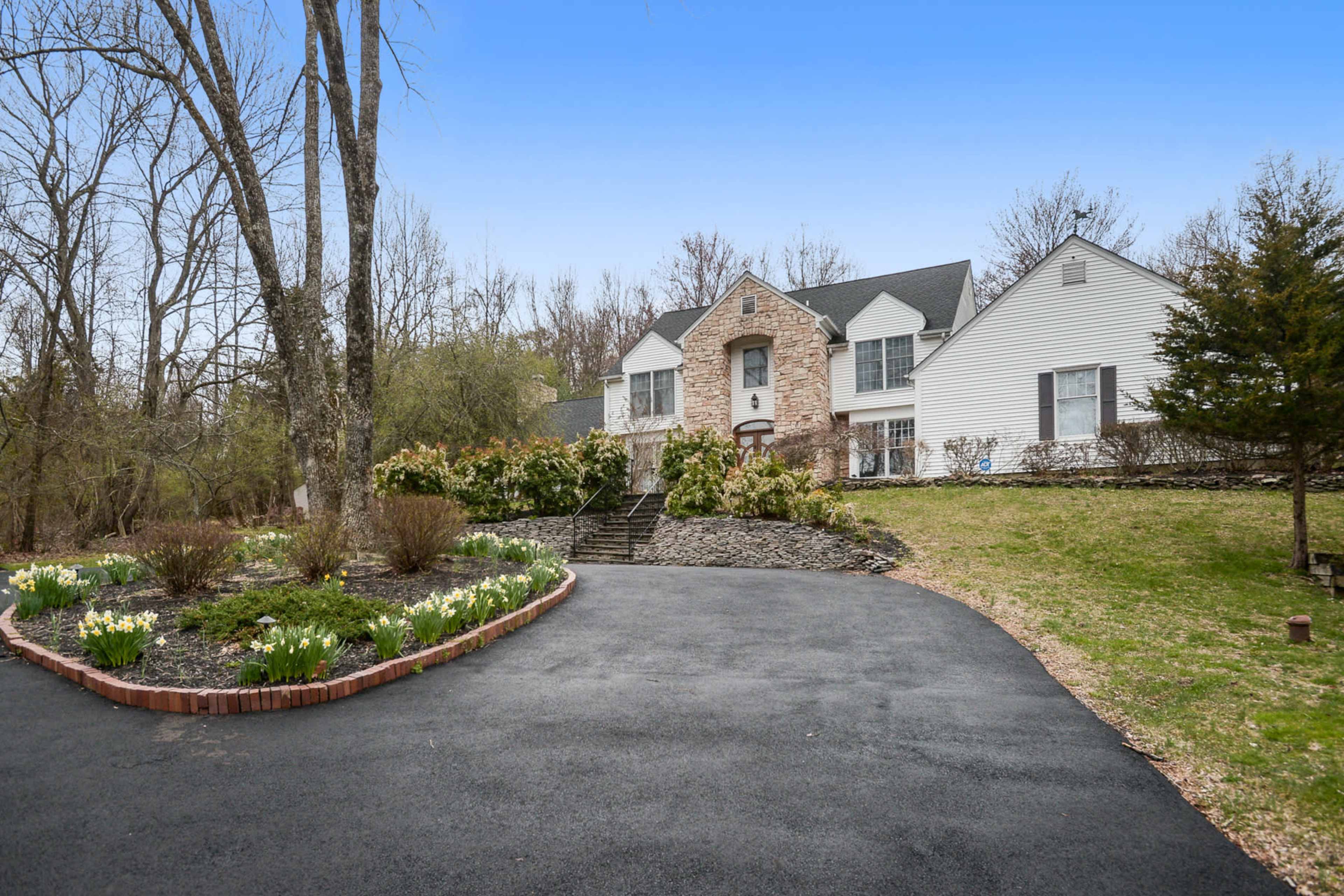 A two-story house with a combination of brick and siding is set back from a winding driveway, surrounded by landscaped gardens and trees.