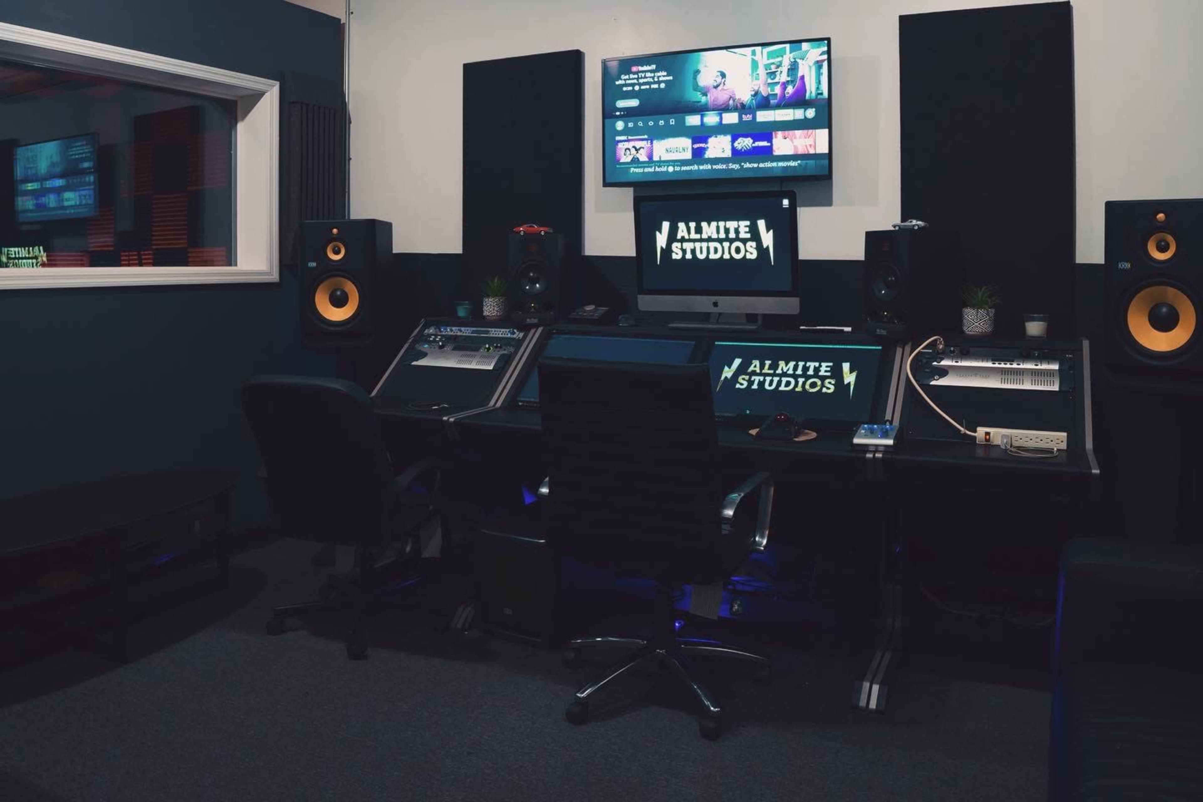 A modern music studio setup with a computer, audio monitors, and sound equipment arranged around a central black desk.