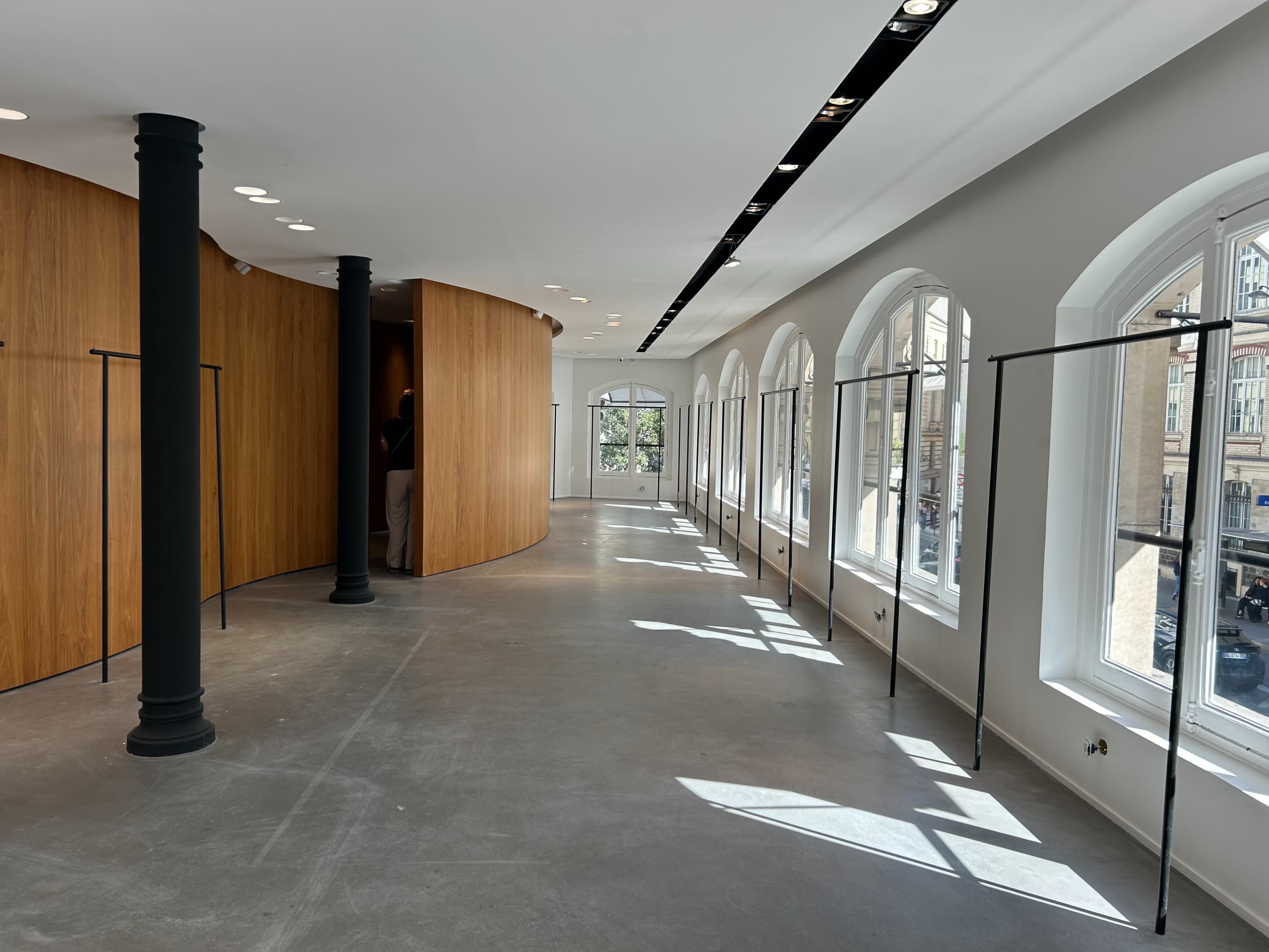 The image shows a modern interior space with curved wooden walls, large windows allowing natural light to enter, and minimalistic clothing racks along the sides.