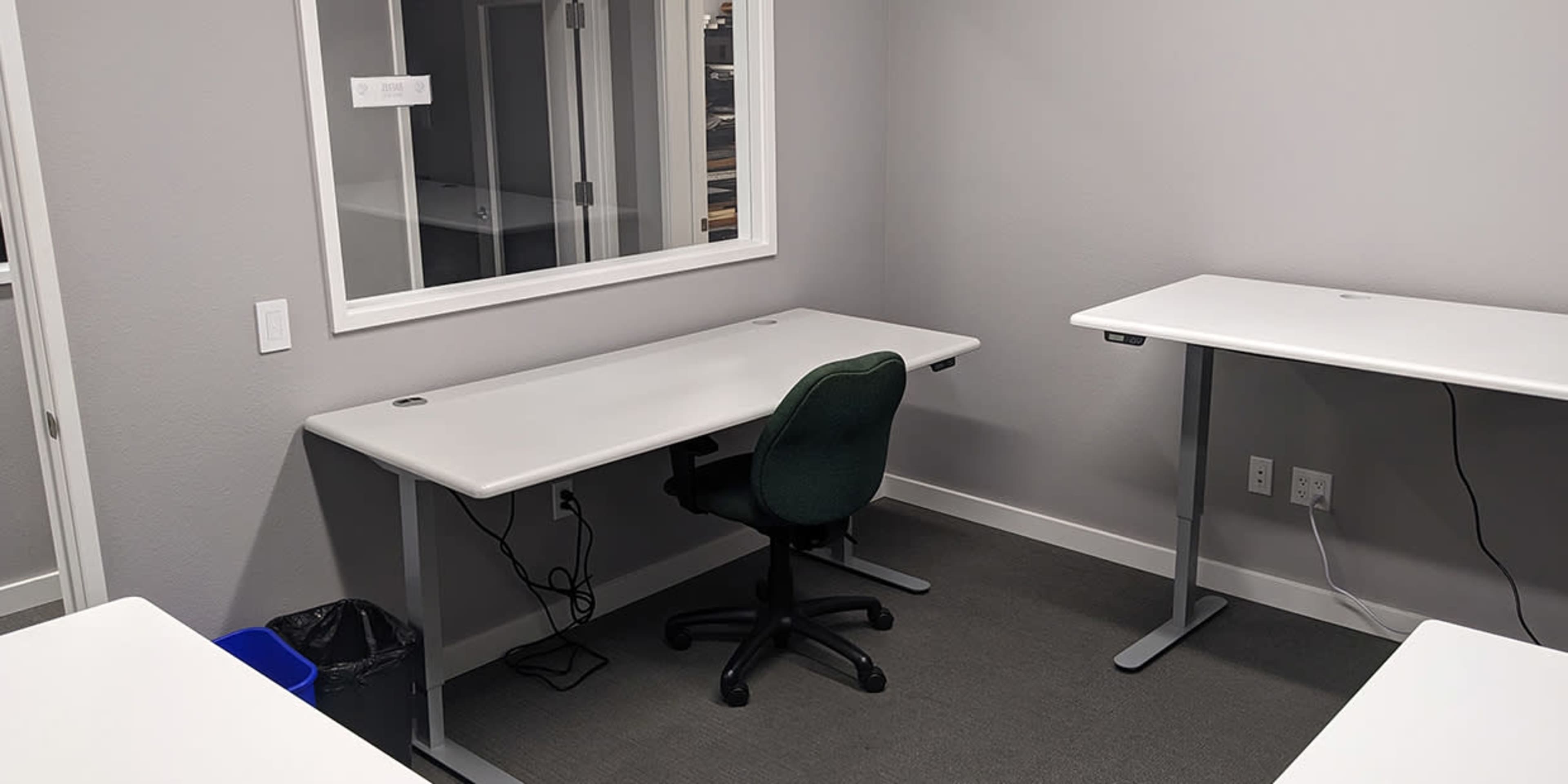 A small, sparsely furnished office with two white desks, a green chair, and a trash bin.