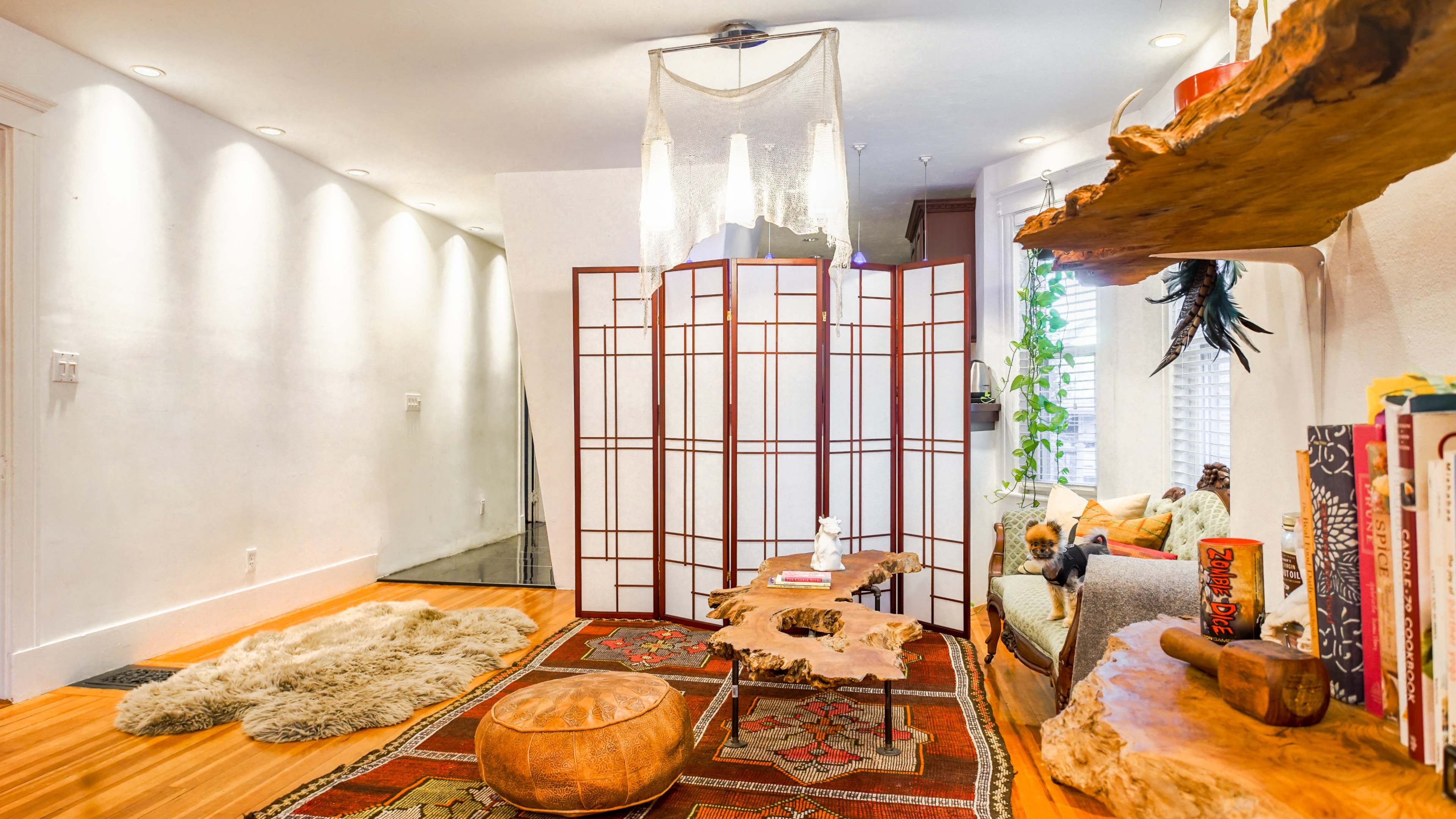 The image features a cozy living area with a wooden coffee table, a plush rug, a folding screen, and various plants, alongside a comfortable seating arrangement.