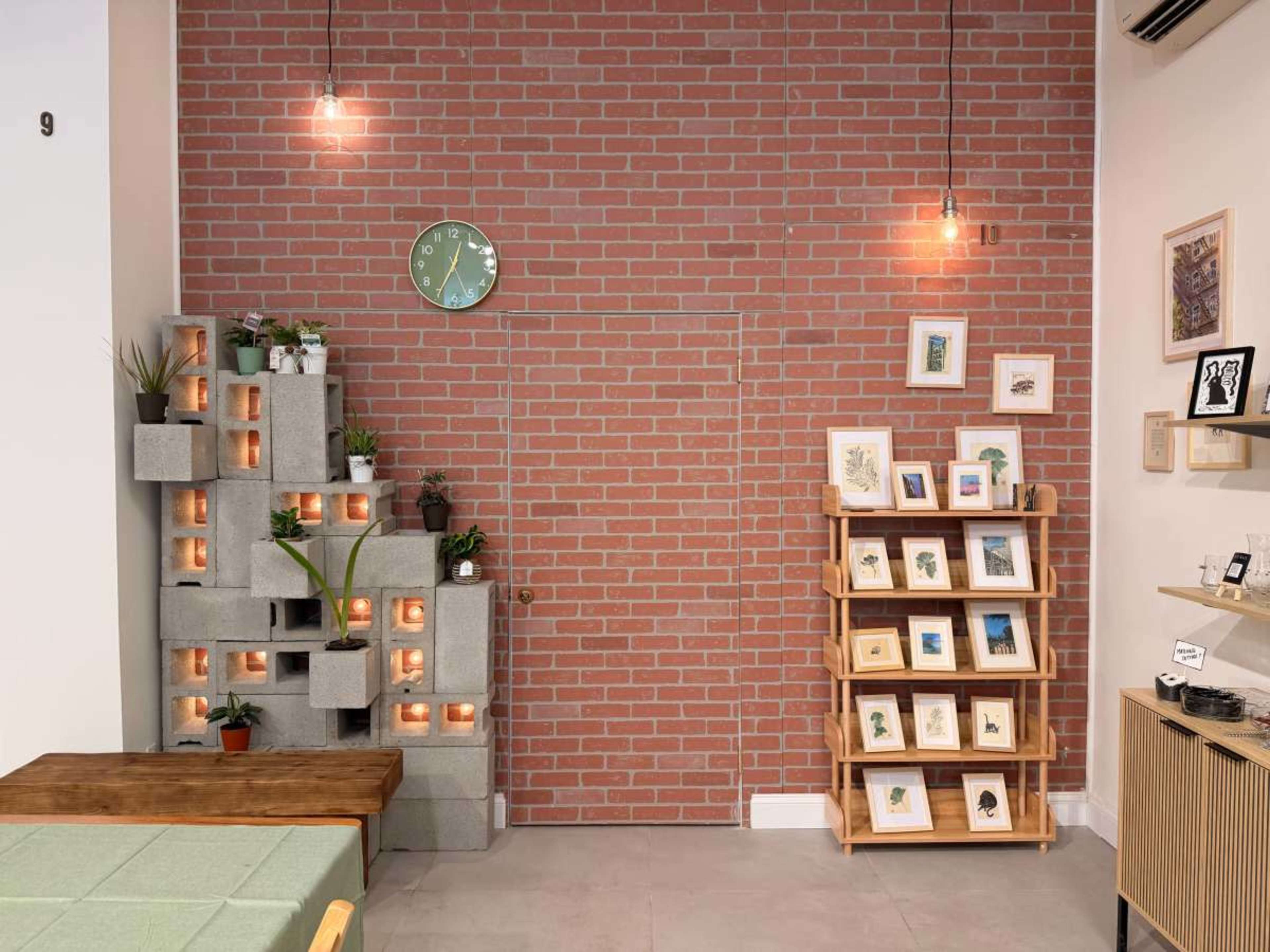 The image shows a cozy cafe interior with a brick wall, decorative plants in concrete blocks, a wall clock, and a display shelf featuring framed art.