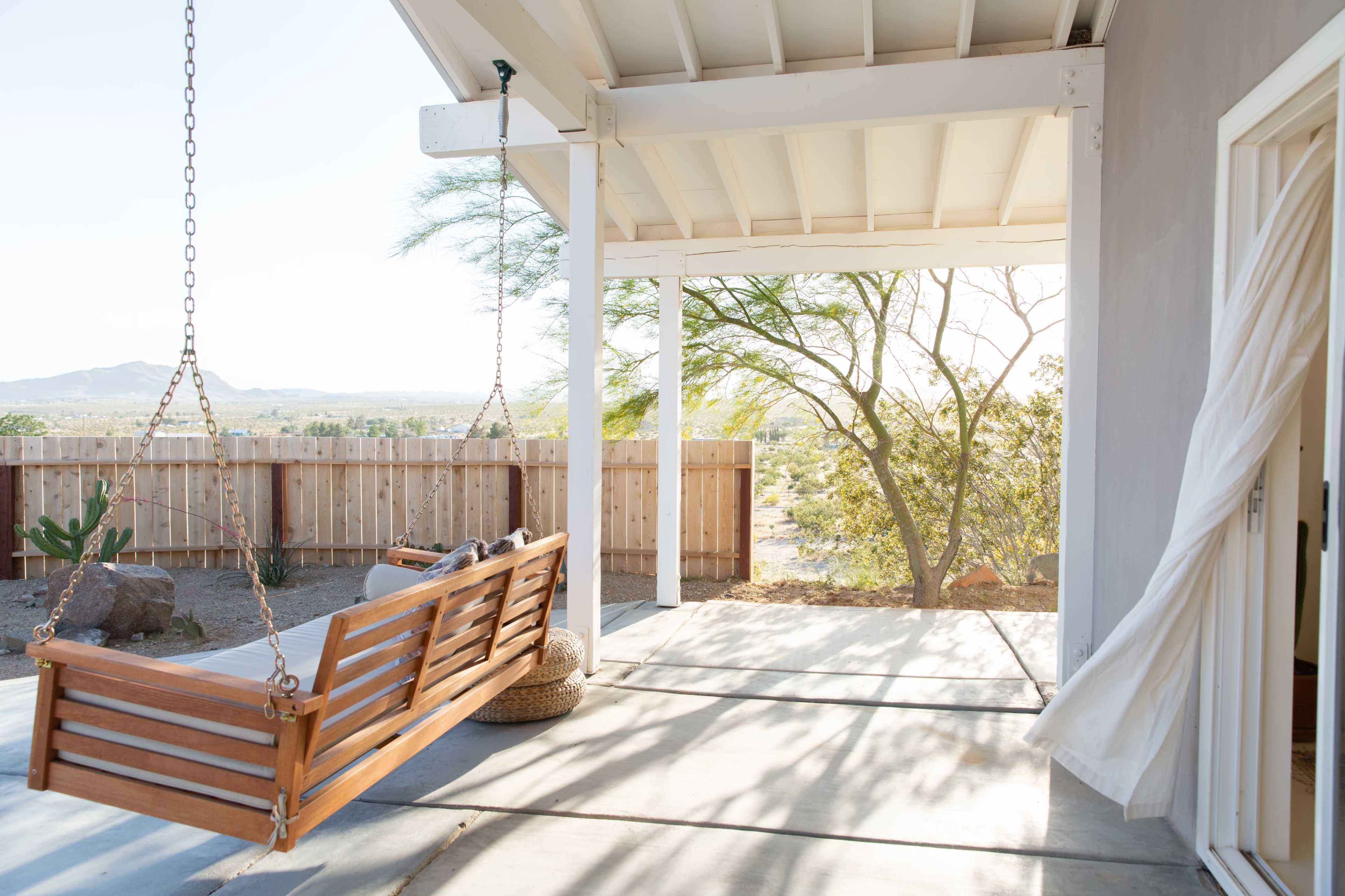 A wooden swing hangs from a covered porch overlooking a desert landscape with distant hills and sparse vegetation.