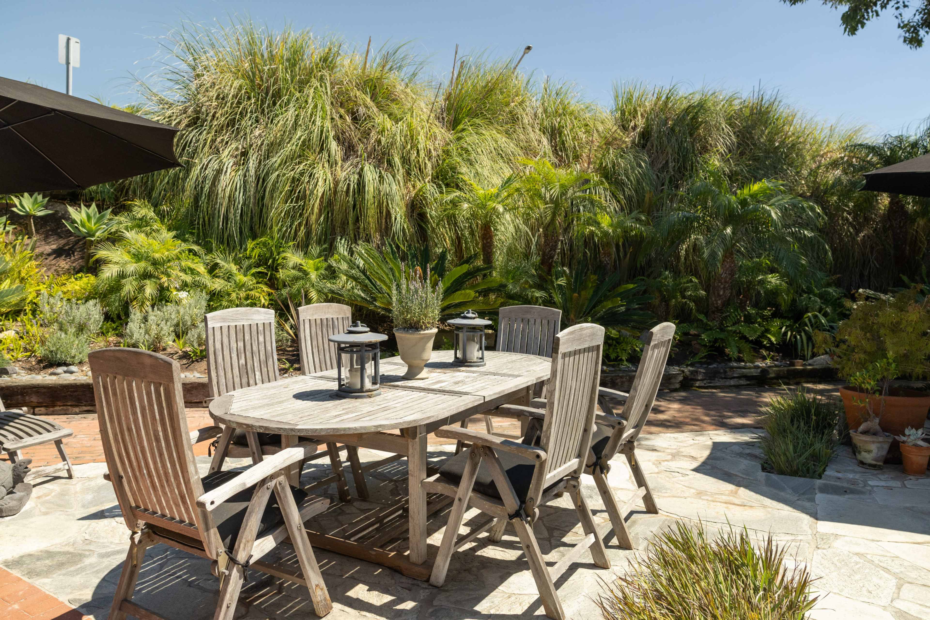 A wooden outdoor dining table with six chairs is surrounded by lush greenery and potted plants in a sunlit garden.