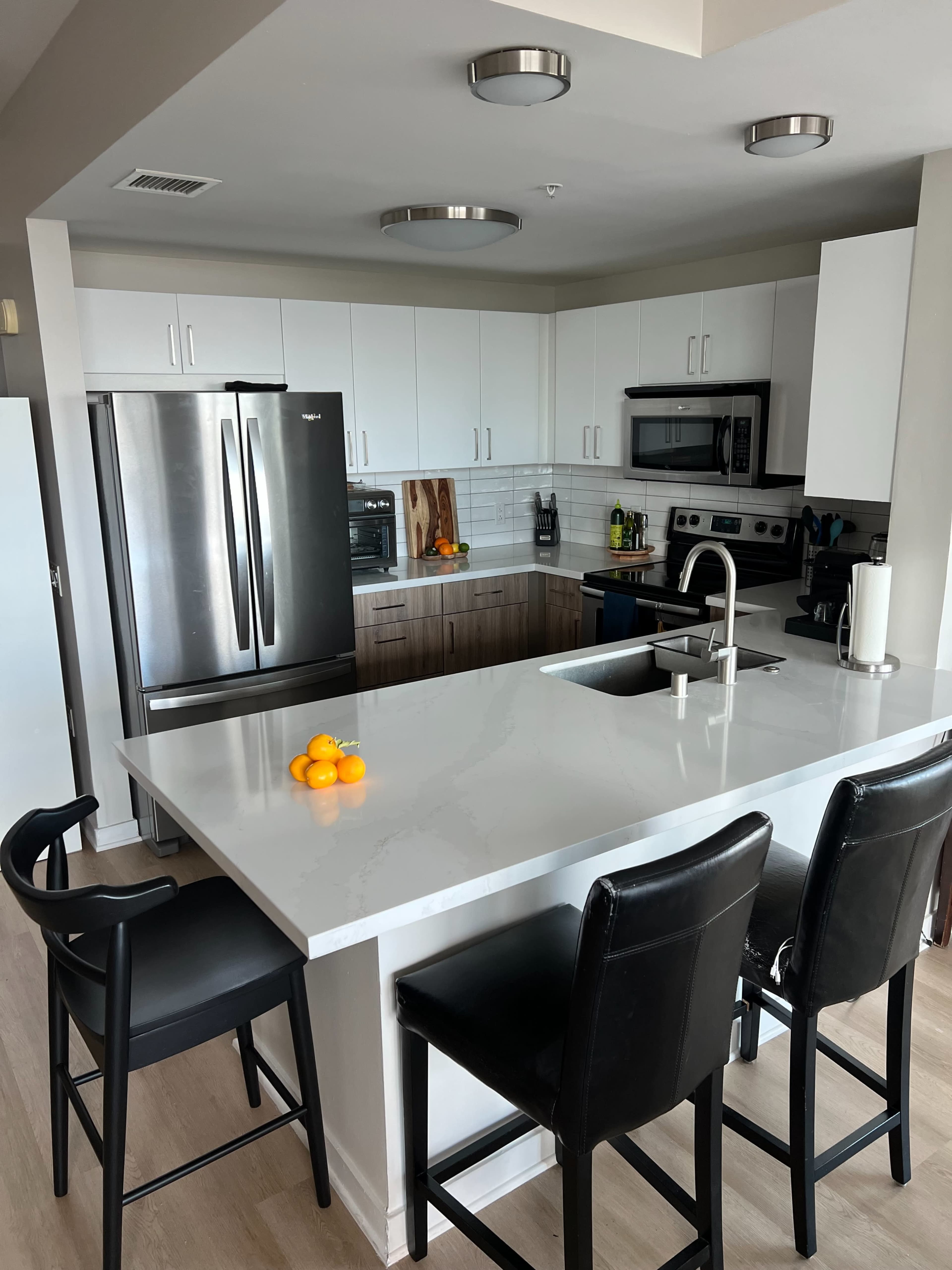 The image shows a modern kitchen with white cabinets, stainless steel appliances, and a central island with three black chairs and a bowl of oranges on the countertop.