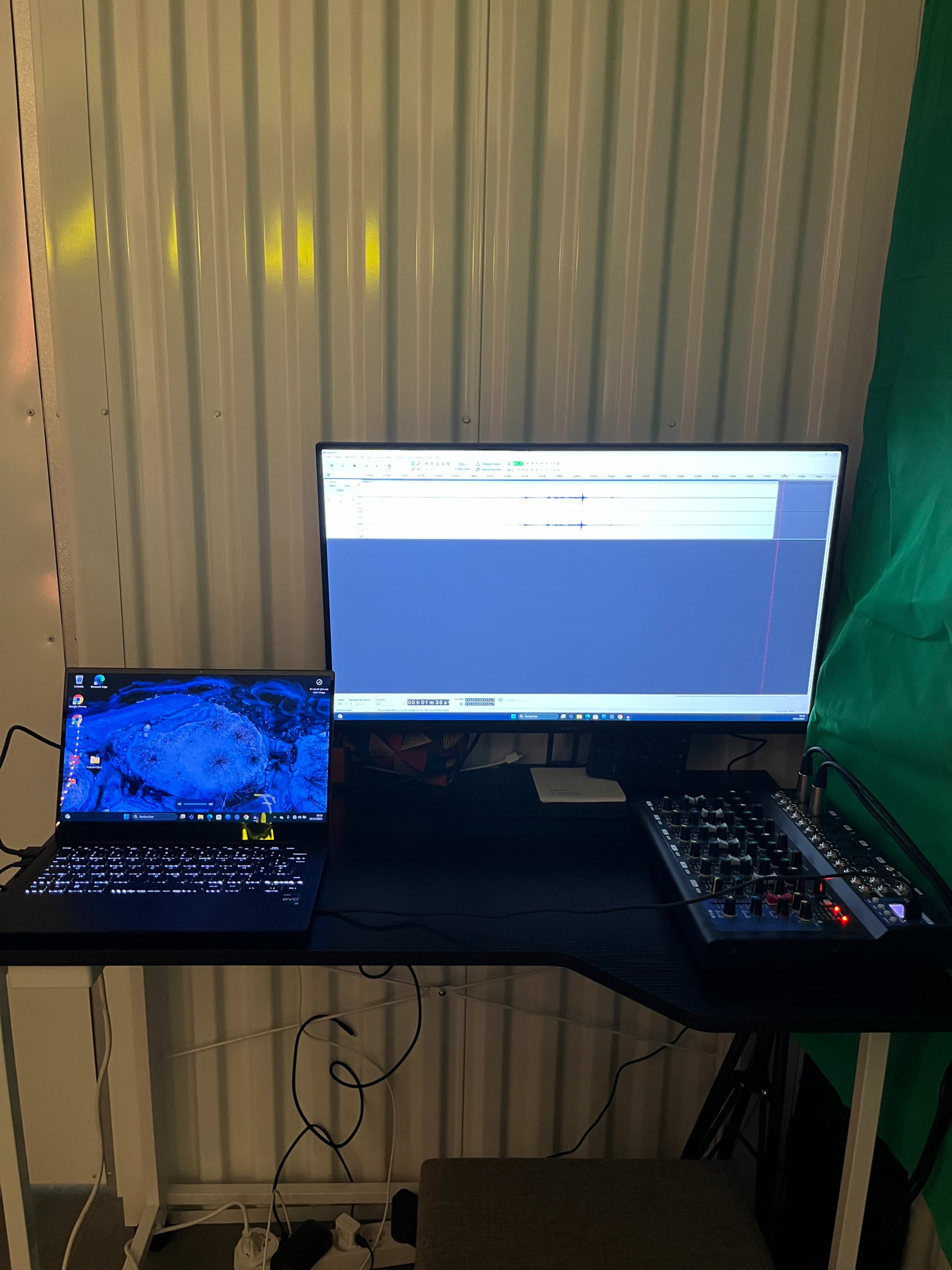 The image shows a workspace with a laptop and a larger monitor displaying audio editing software, alongside an audio mixer on a black table.