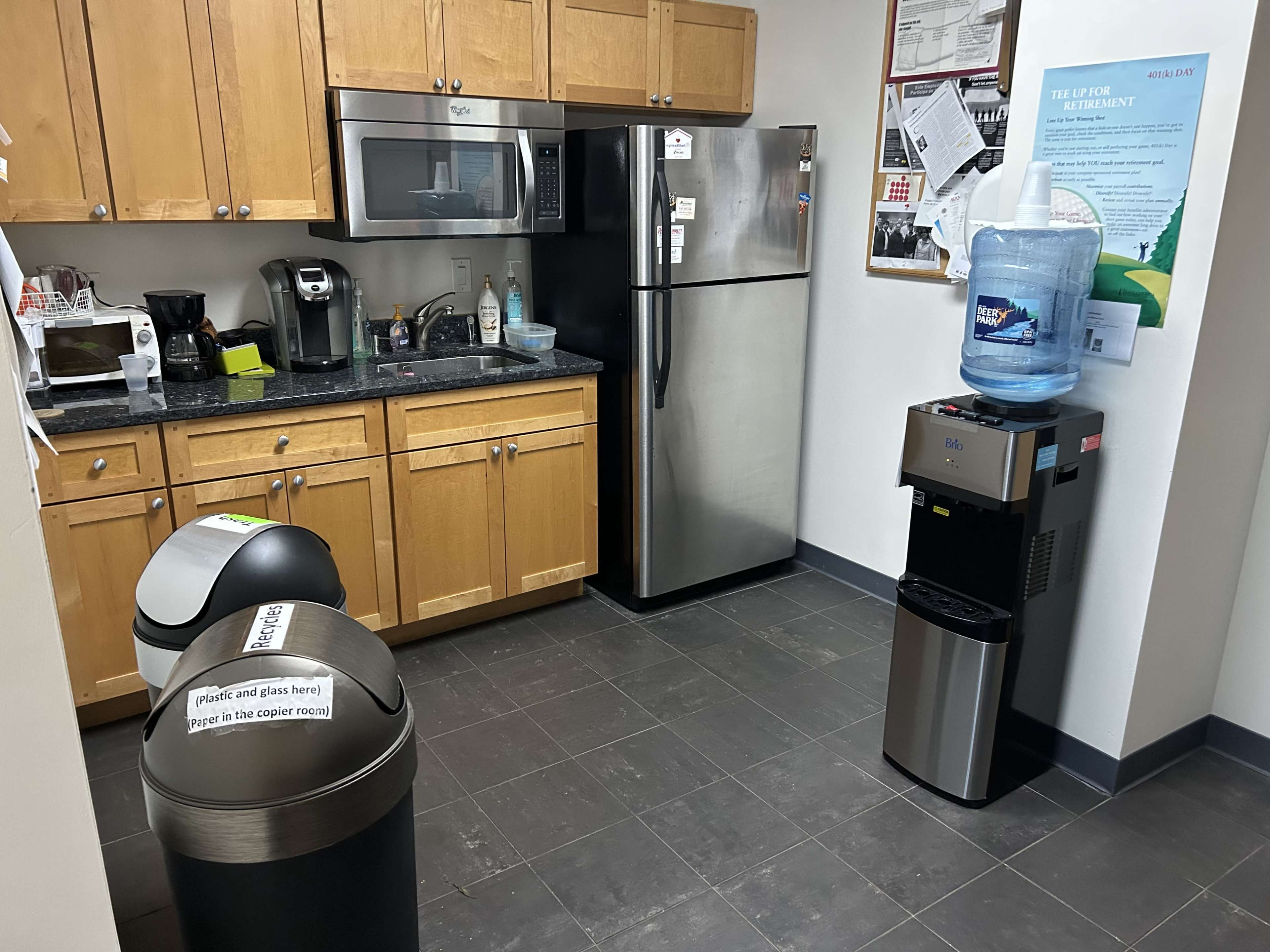 A kitchen area with wooden cabinets, a microwave, a stainless steel refrigerator, and a water cooler next to two trash bins.