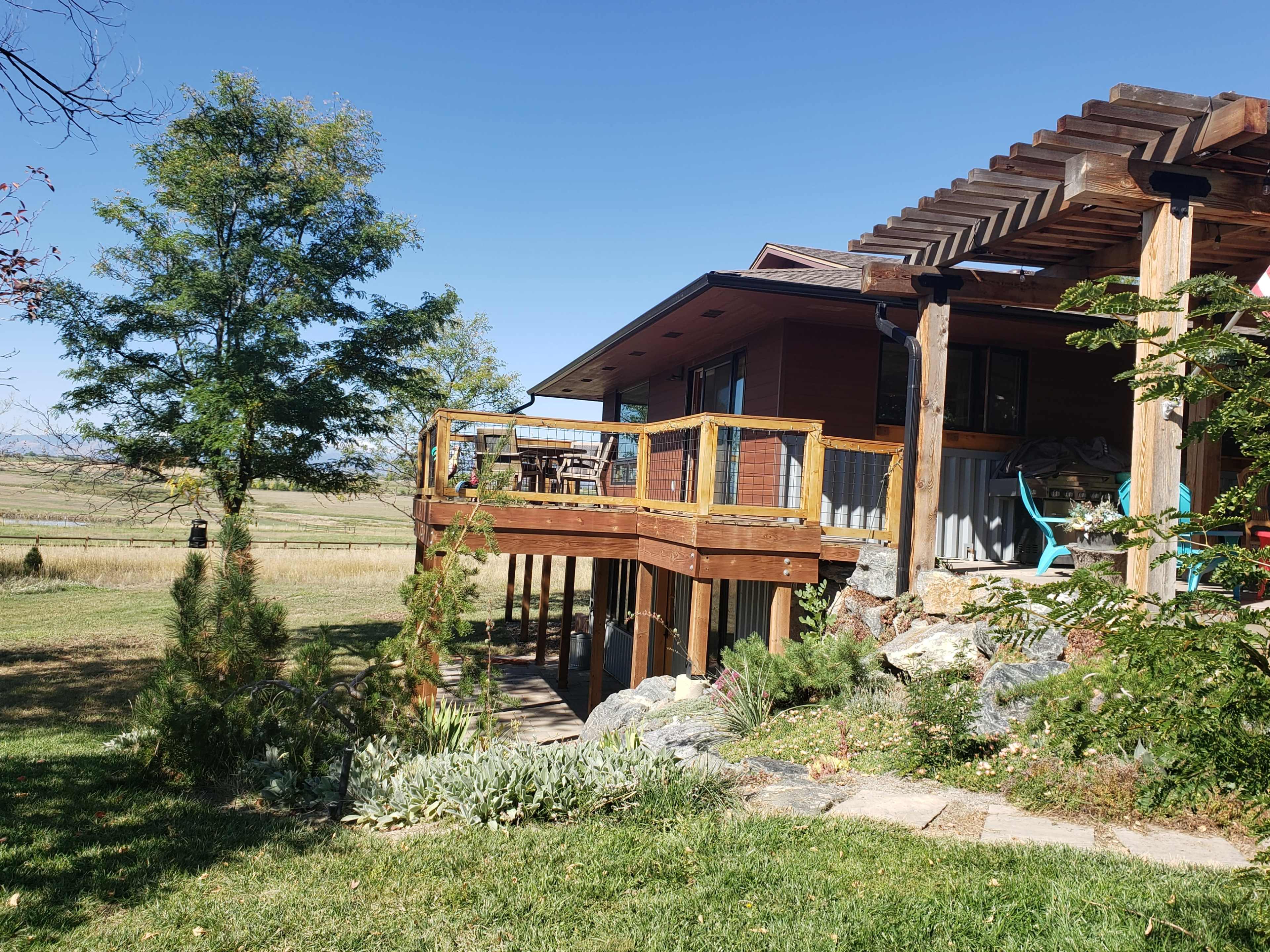 A wooden house with a balcony overlooks a grassy area and is surrounded by trees and landscaping.