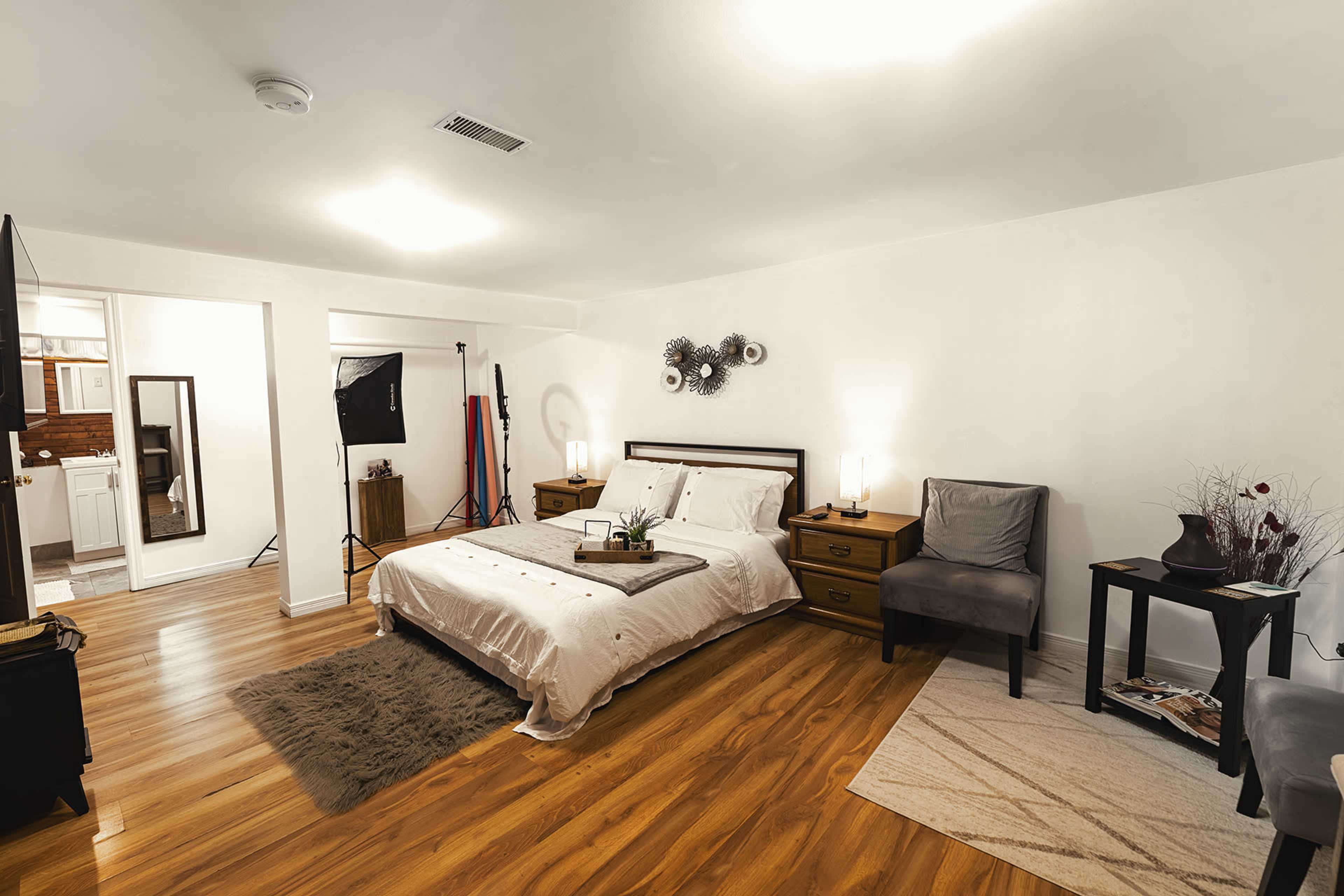 A well-lit bedroom featuring a bed with white linens, two nightstands, a chair, and a small table, all set on a hardwood floor.