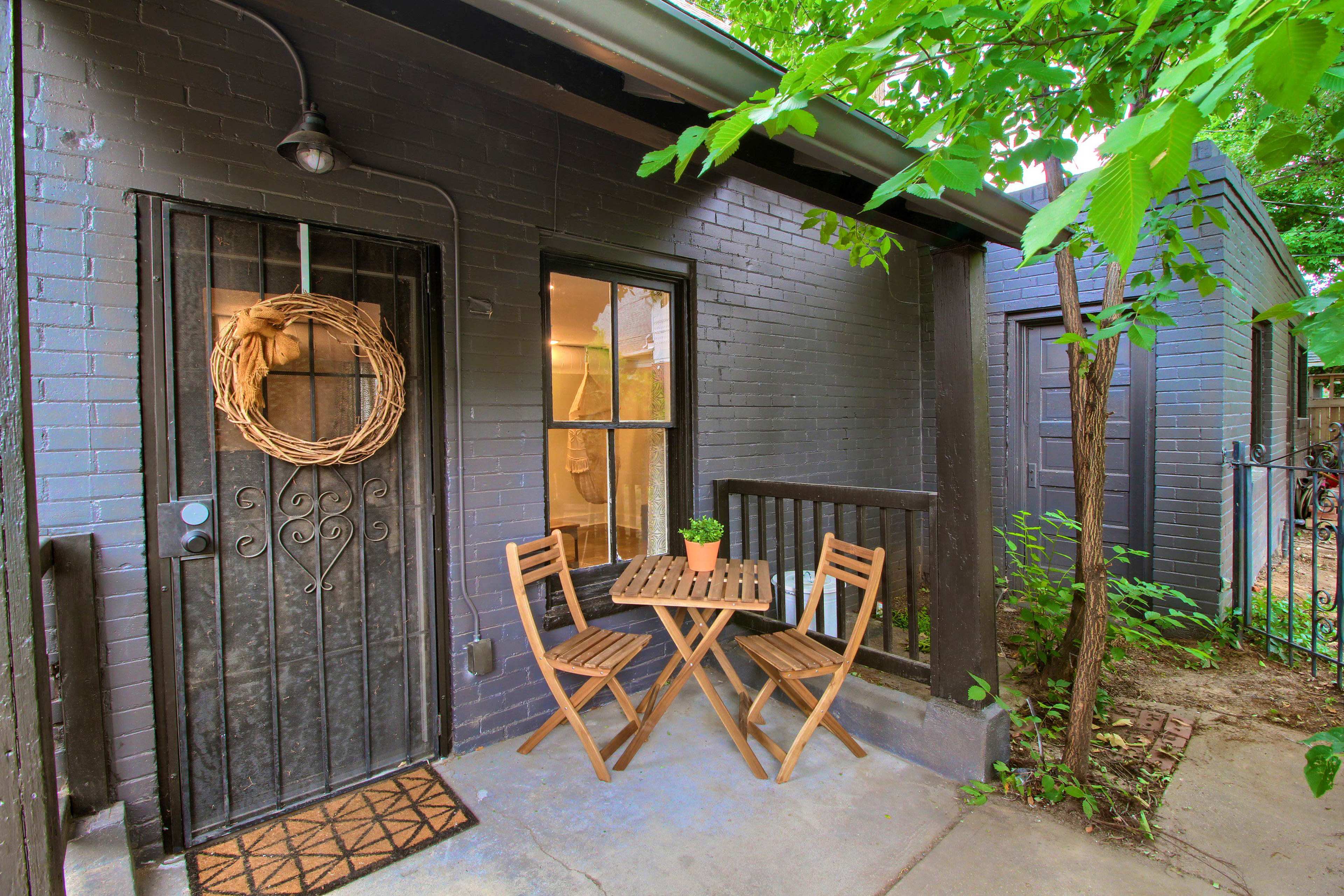 A small outdoor seating area features a wooden table and two folding chairs in front of a dark brick building with a decorative wreath on the door.