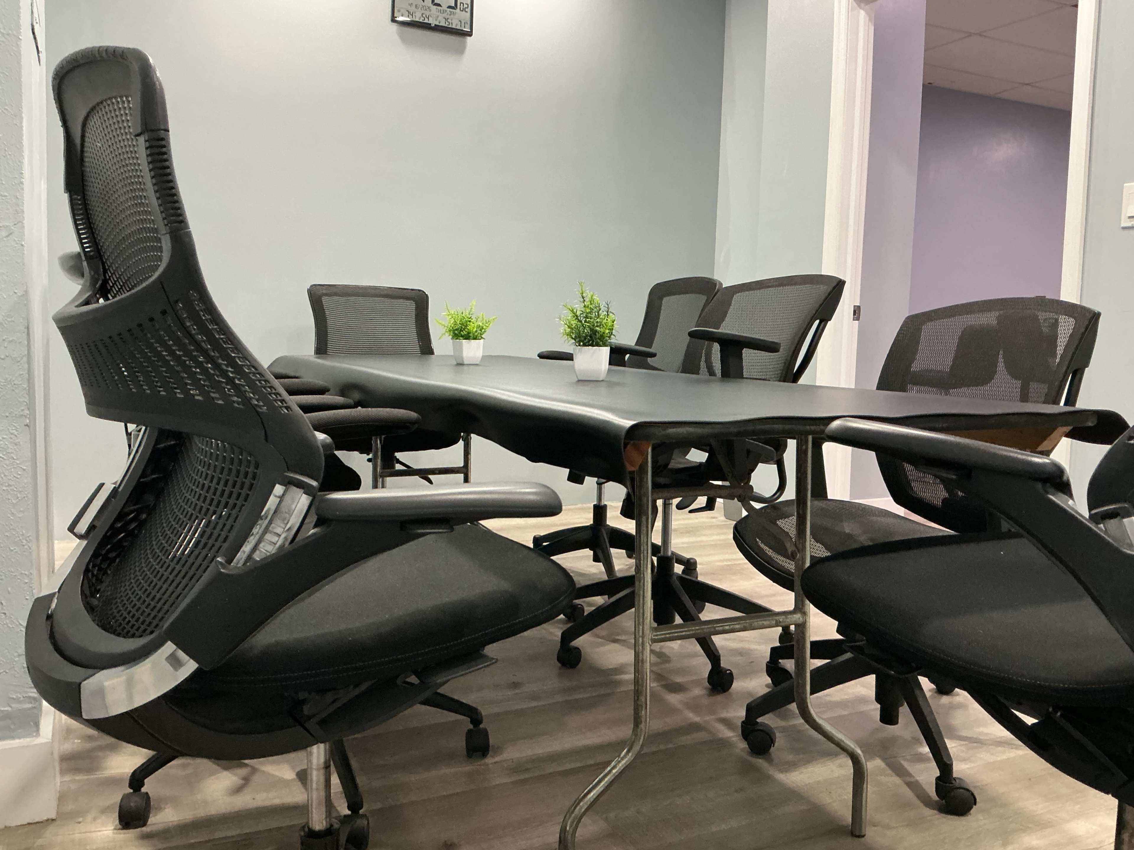 A conference room features a large rectangular table surrounded by several black ergonomic chairs and small potted plants.
