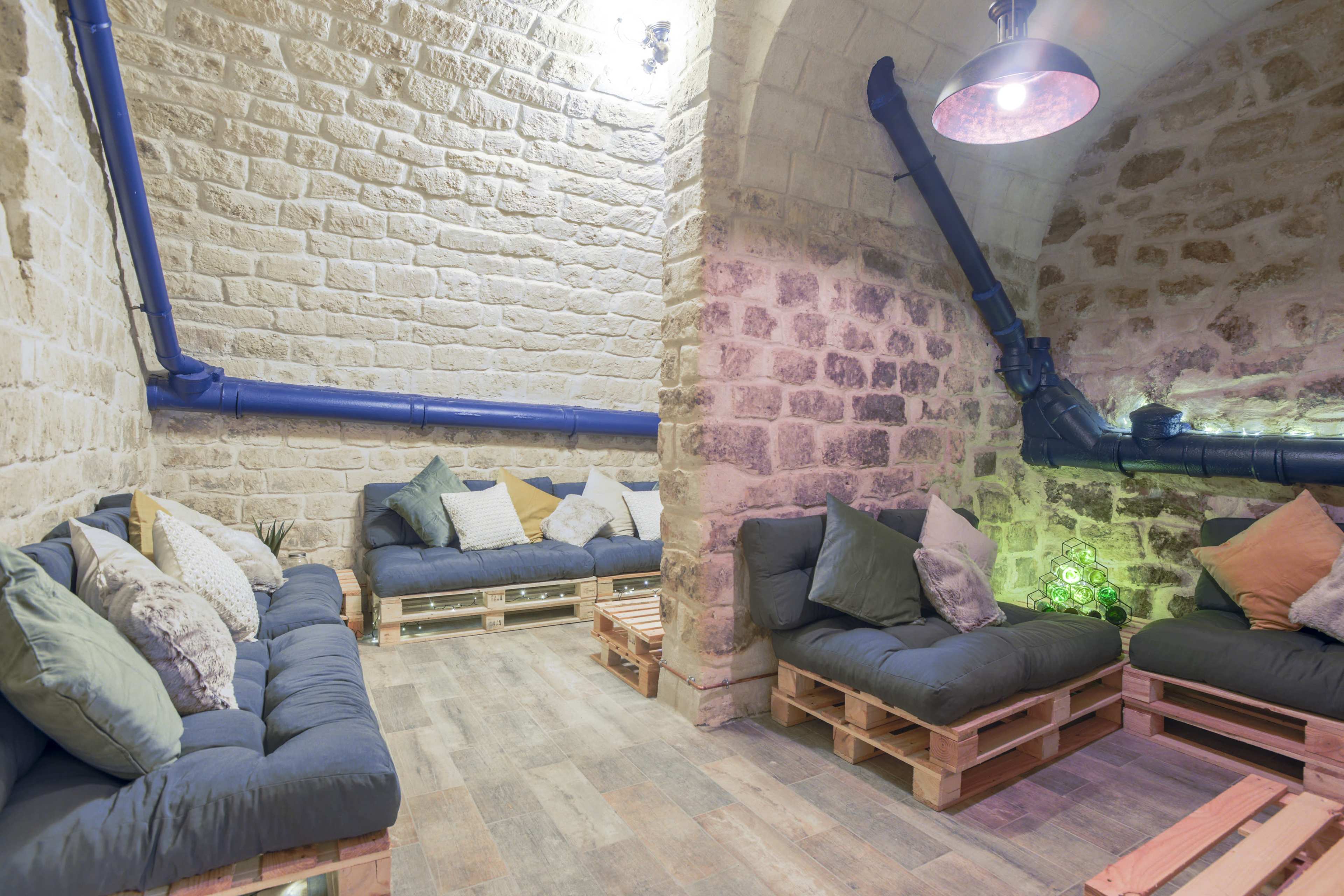 A cozy lounge area with pallet seating and decorative pillows, set against textured stone walls and illuminated by soft lighting.