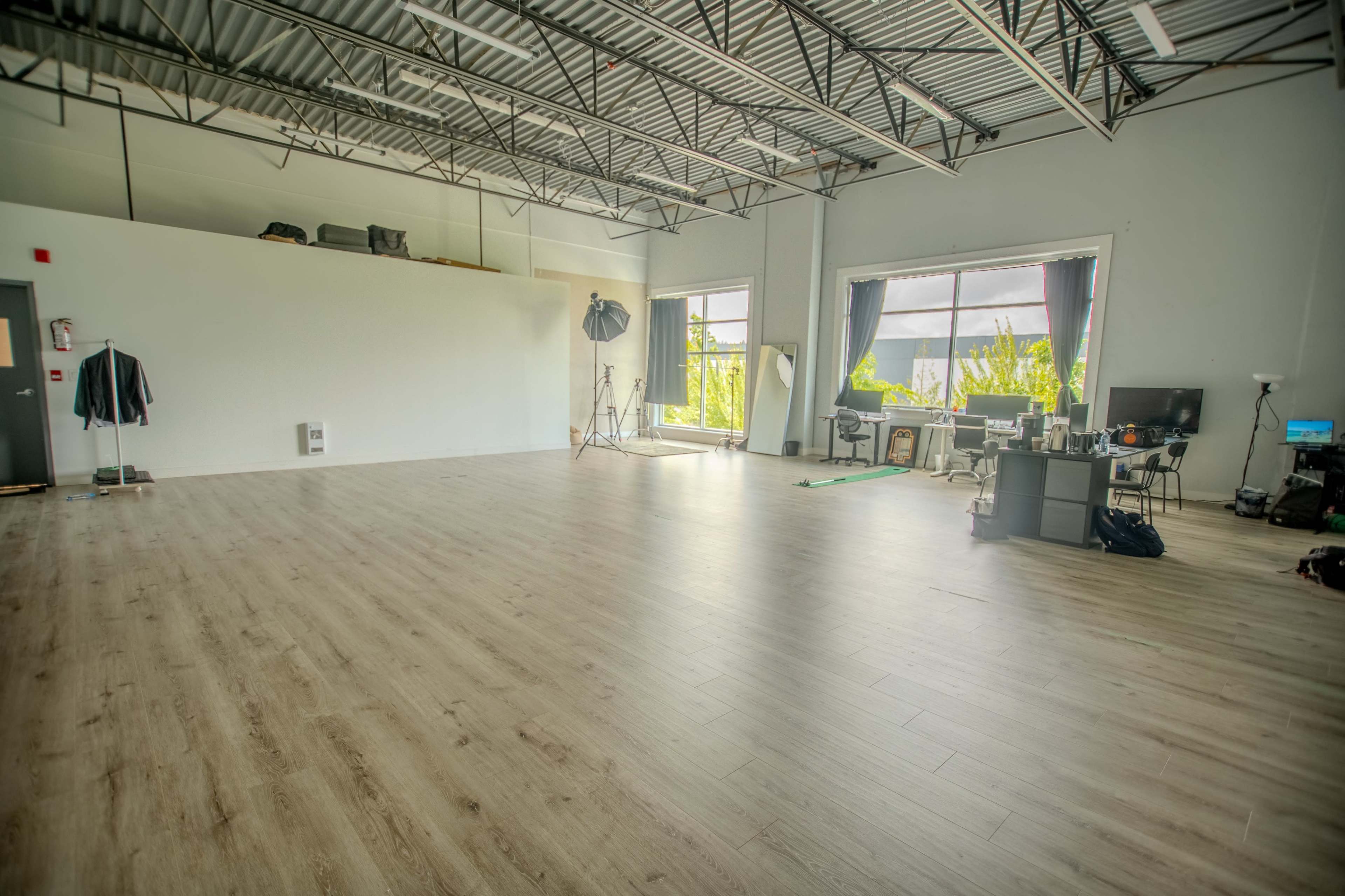 The image shows a spacious studio with wooden flooring, tall ceilings, and large windows, featuring various photography equipment and a work desk in one corner.
