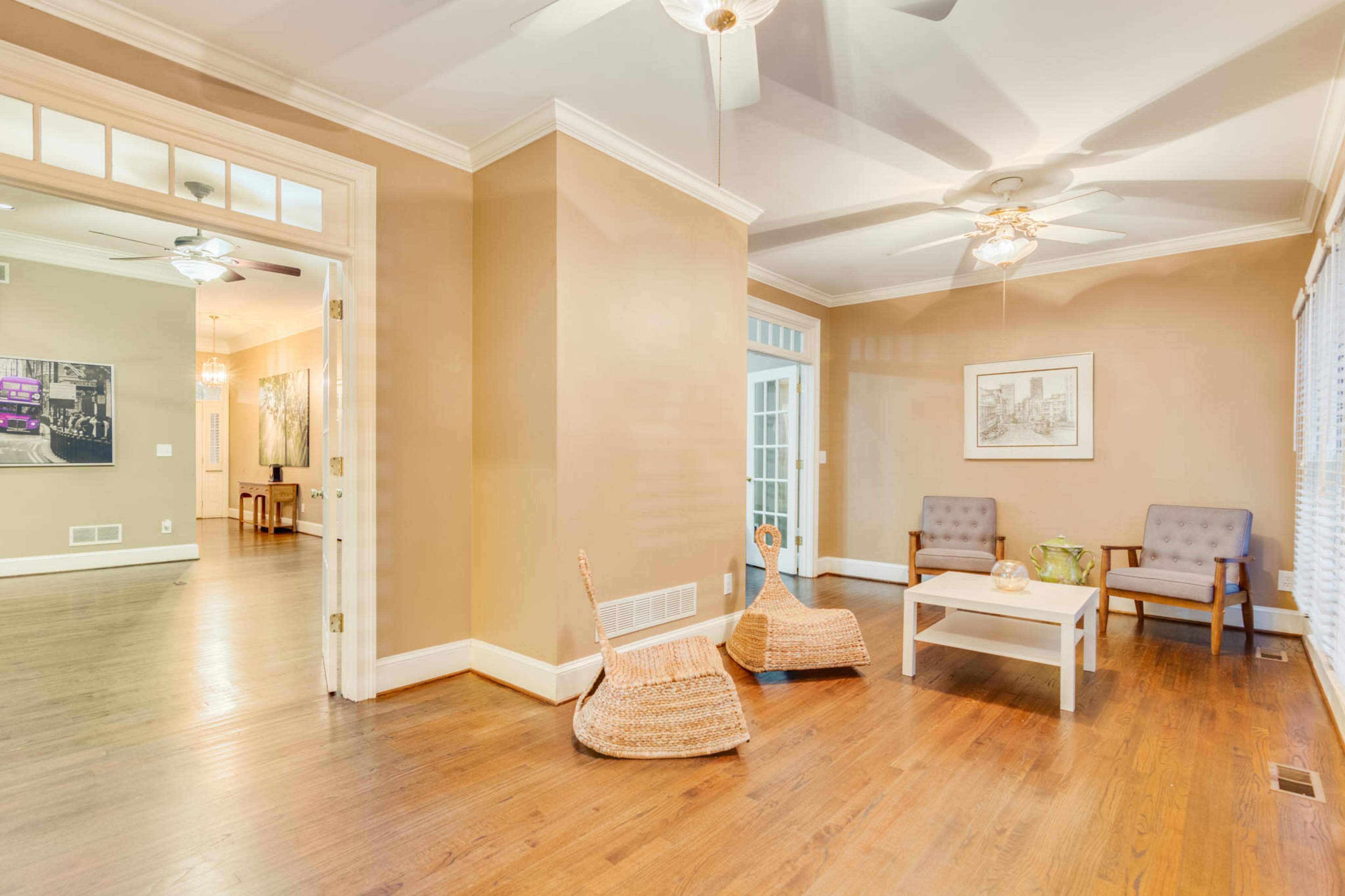 The image shows a well-lit living area with two chairs, a small coffee table, and wooden floors, adjacent to another room.