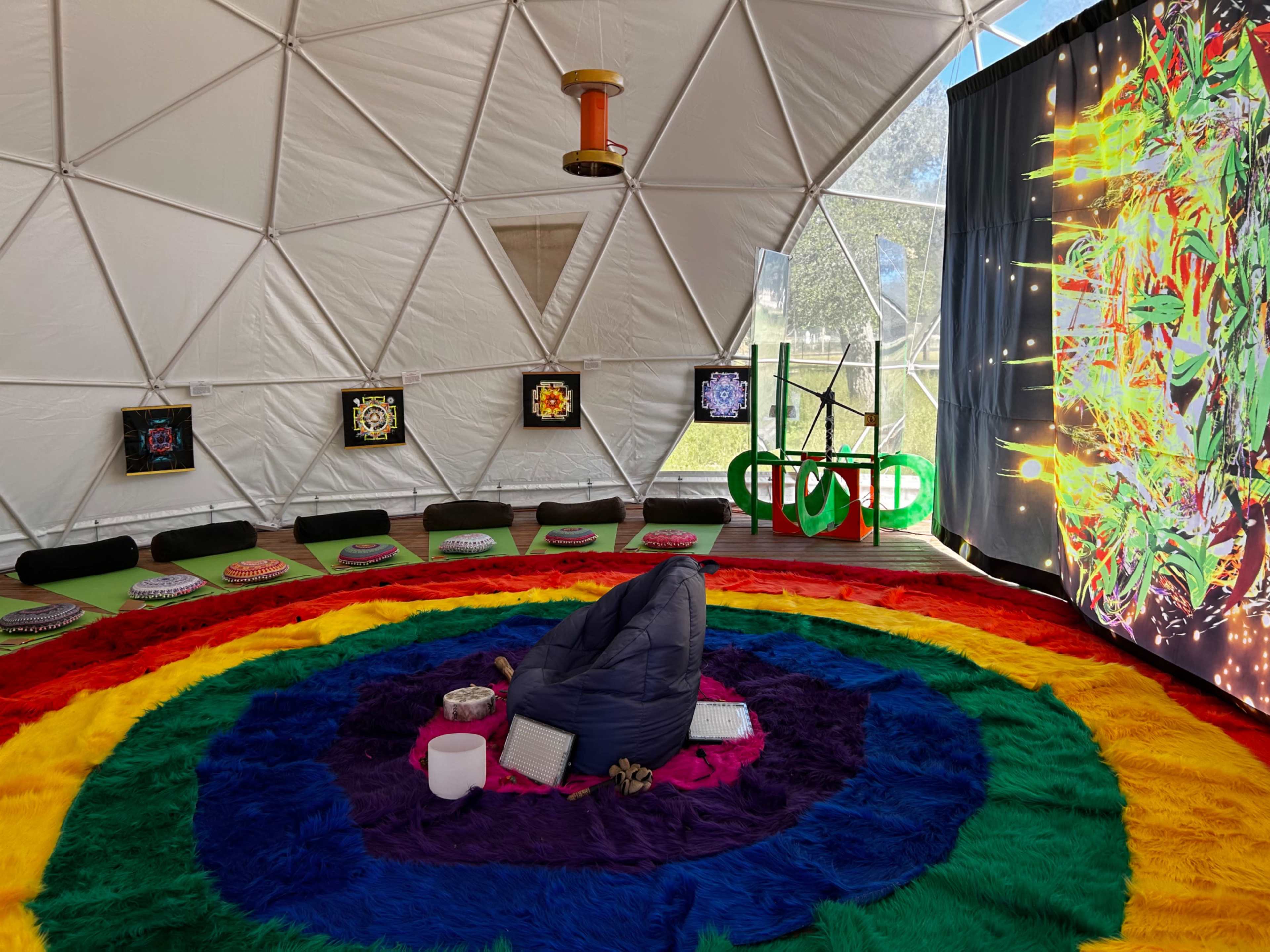 The interior of a geodesic dome features a colorful circular rug, seating around the perimeter, and vibrant artwork displayed on the walls.