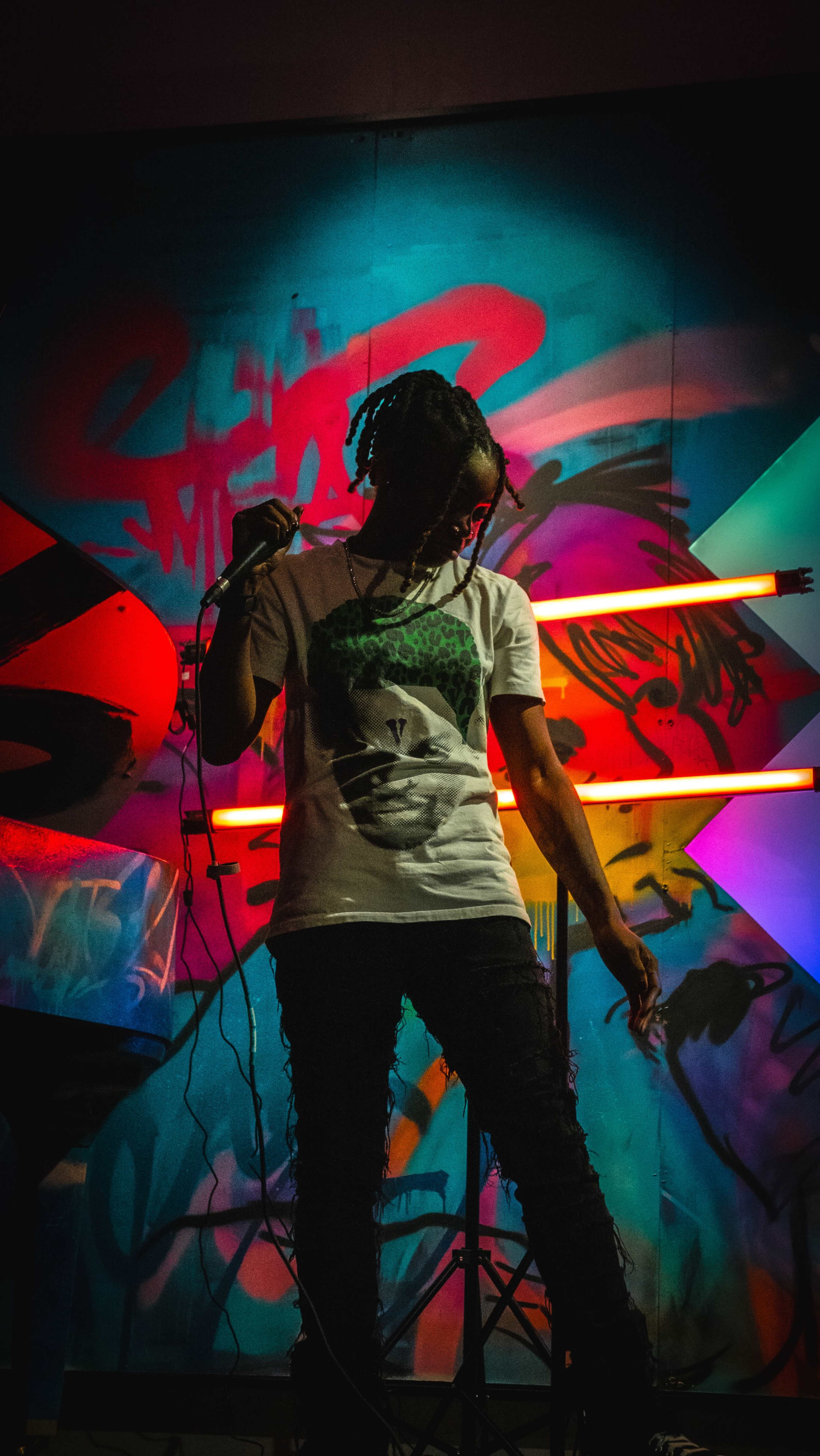A person stands on stage holding a microphone in front of a colorful, graffiti-covered backdrop with neon lights.