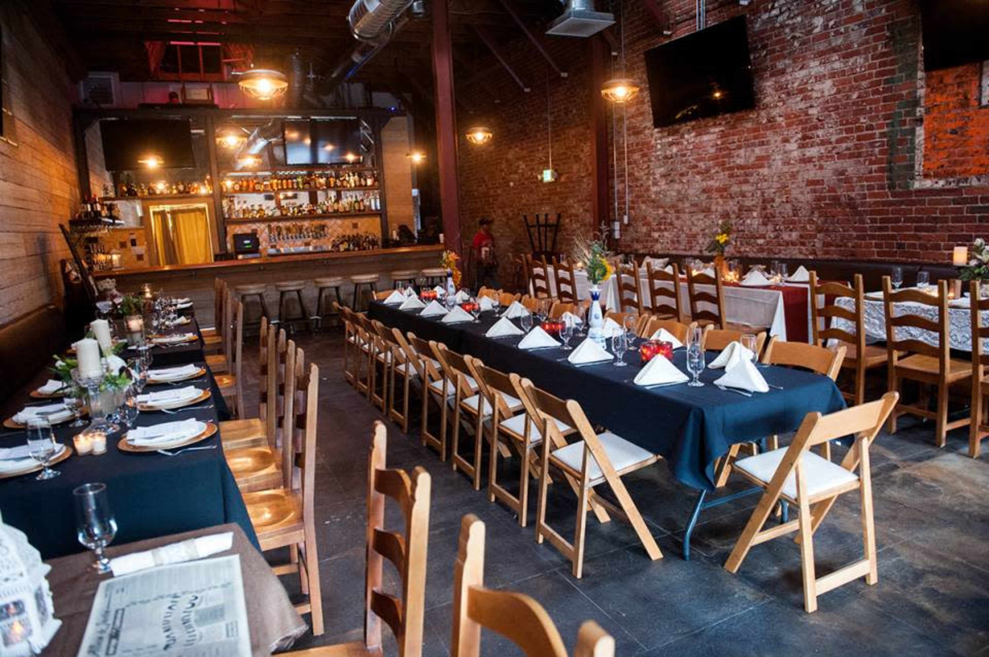 A restaurant interior with a long set of tables arranged for dining, featuring wooden chairs and a bar at the back, against a backdrop of exposed brick walls.
