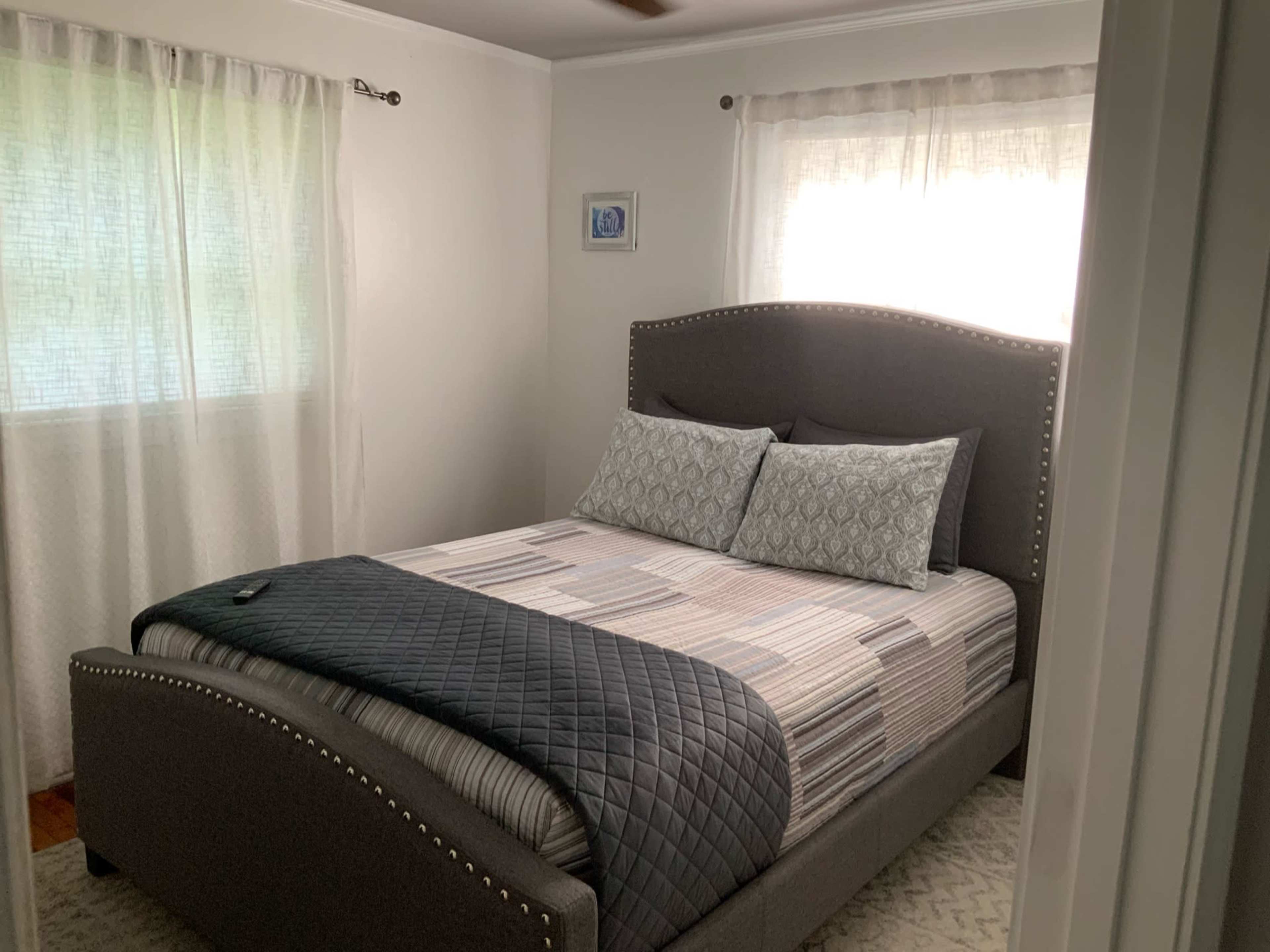 A neatly arranged bedroom with a gray upholstered bed, two pillows, and large windows with sheer curtains.