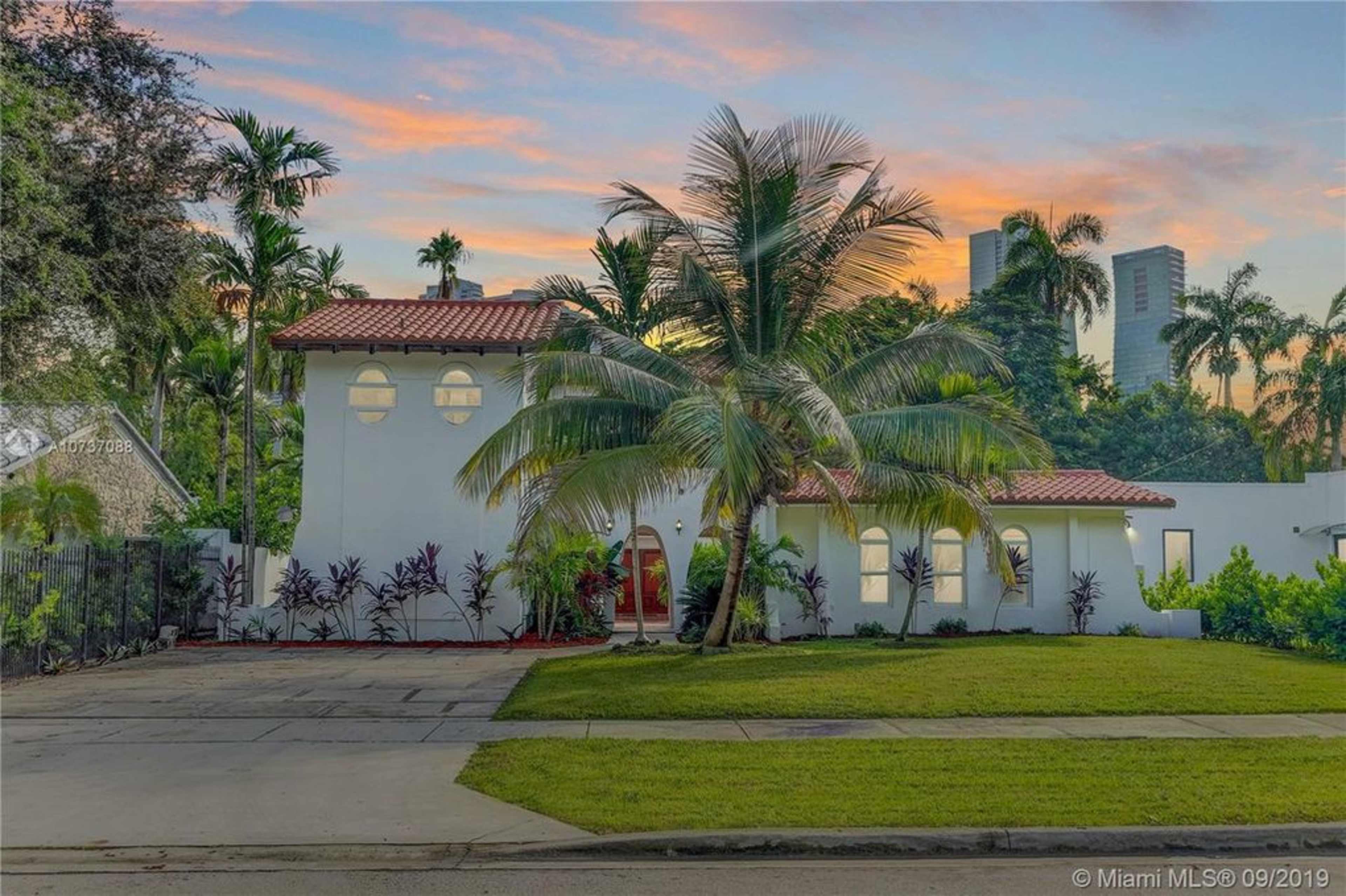 A single-story white house with a red tile roof is surrounded by palm trees and lush greenery, set against a colorful sunset sky.