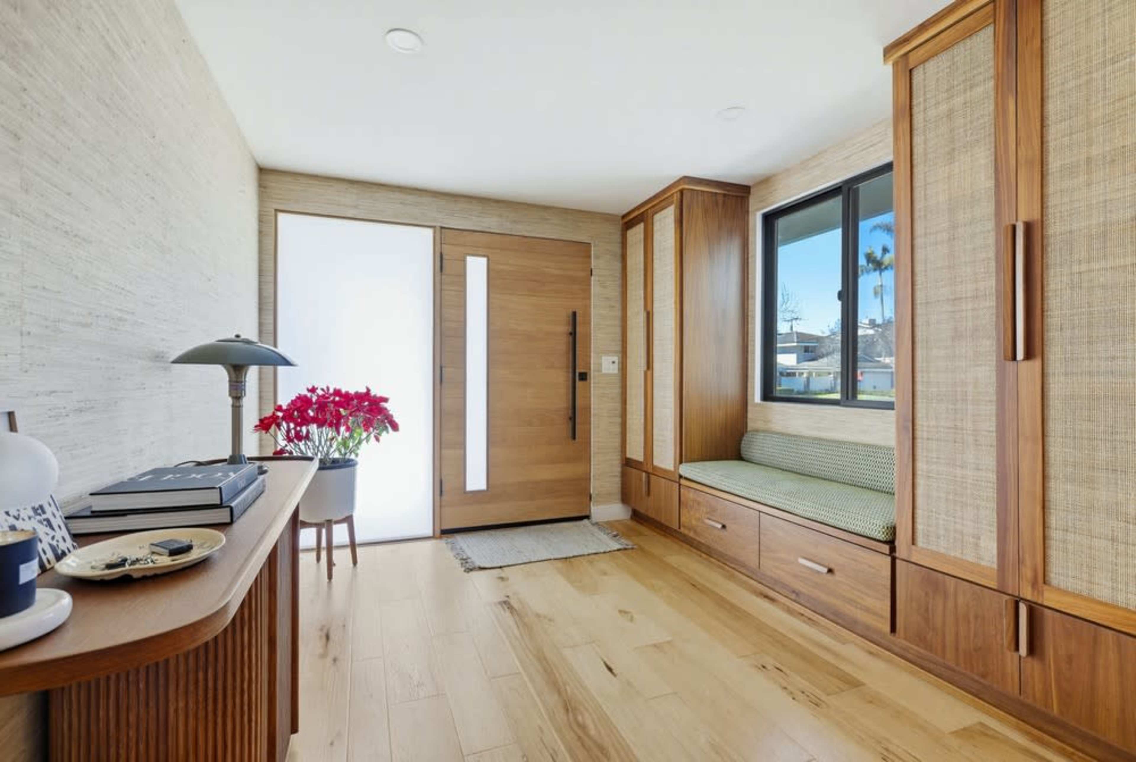 The image shows a modern entryway featuring a wooden door, a window, a built-in bench, and a decorative table with potted flowers.