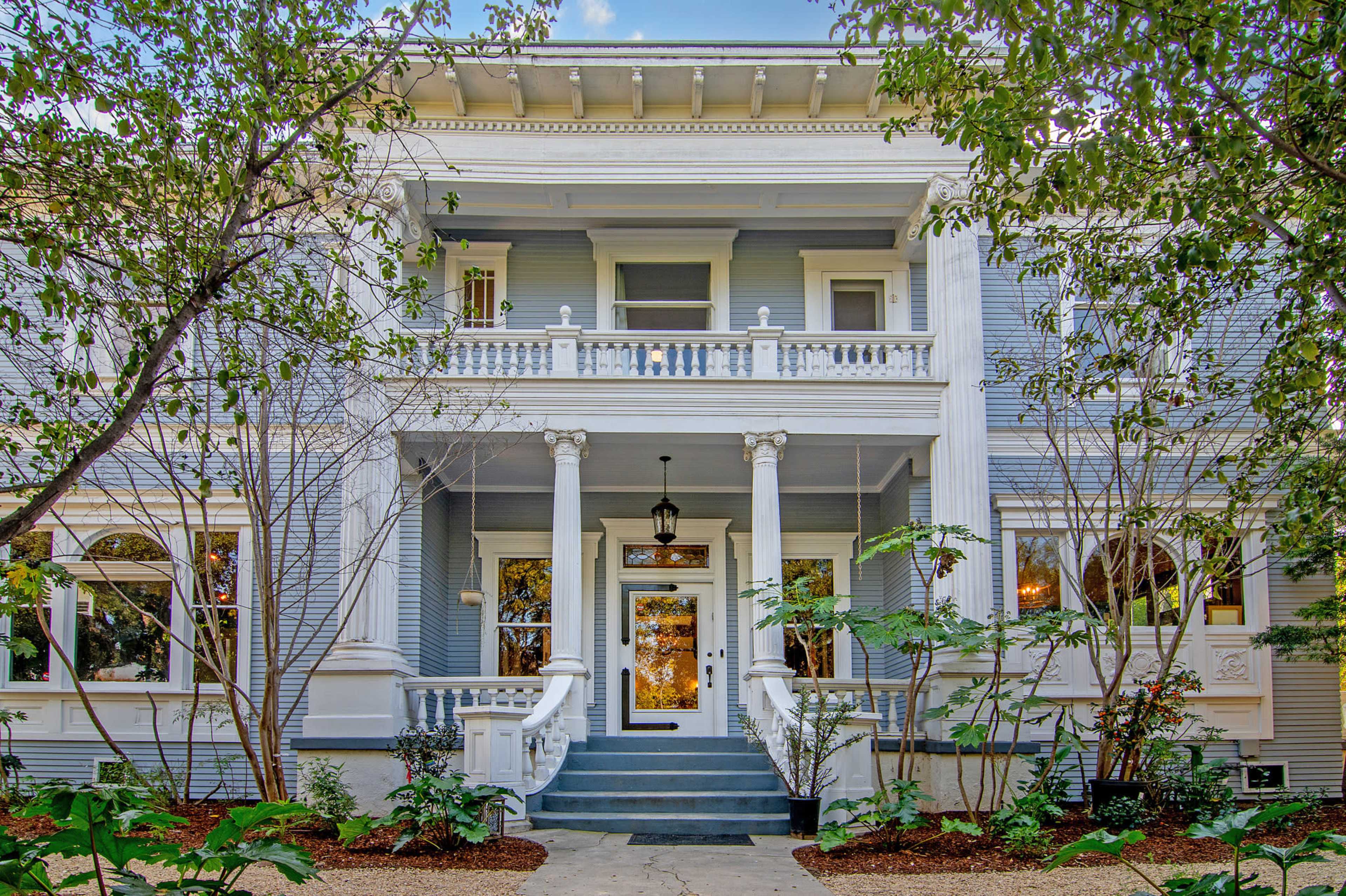 The image features a large, two-story Victorian-style house with a wide front porch supported by tall columns, surrounded by well-maintained greenery and landscaping.