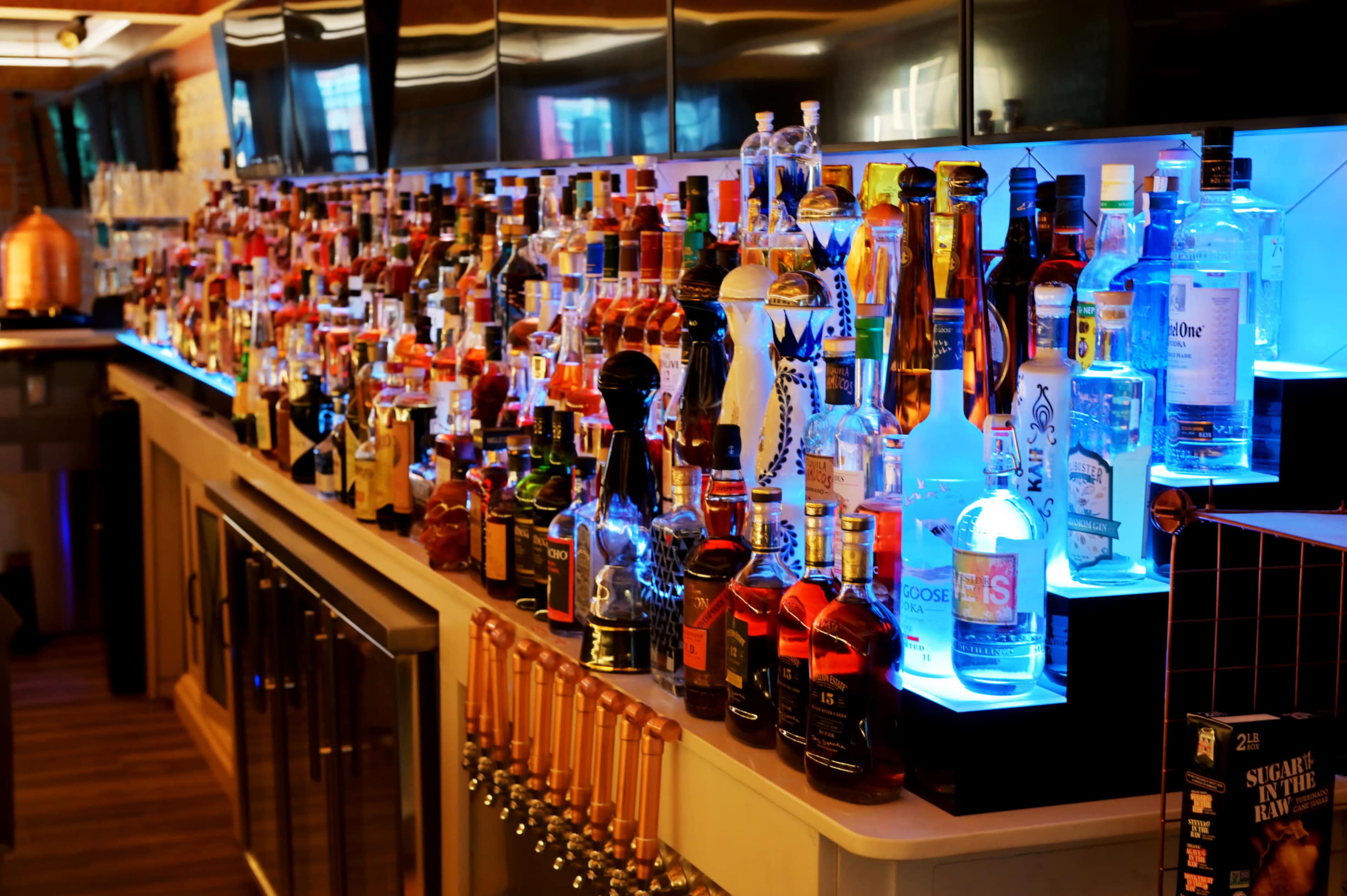 The image shows a bar displaying a wide variety of alcoholic beverages, illuminated by blue lighting.