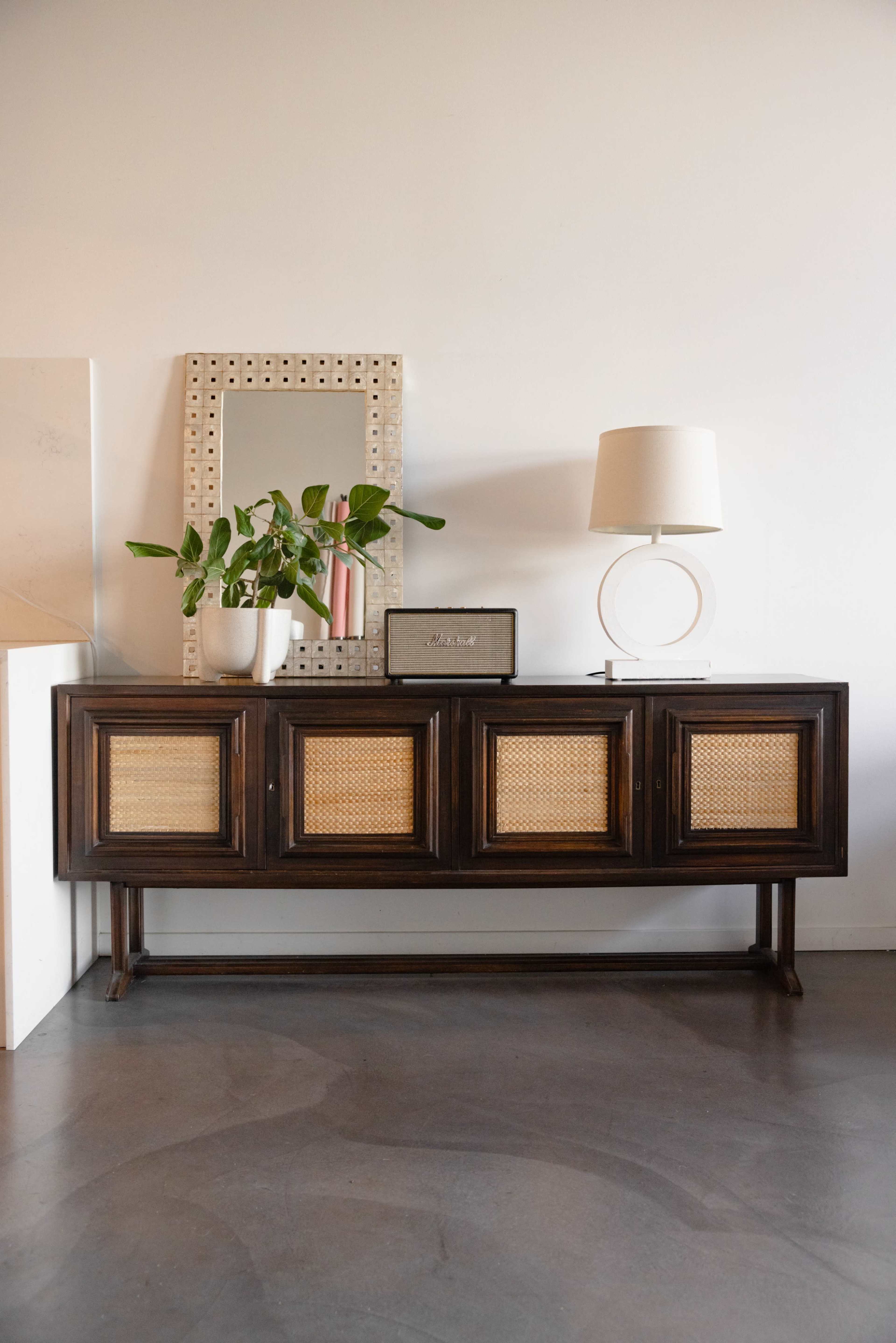 A wooden sideboard with cane paneling is placed against a white wall, accompanied by a lamp, a small mirror, and a potted plant.