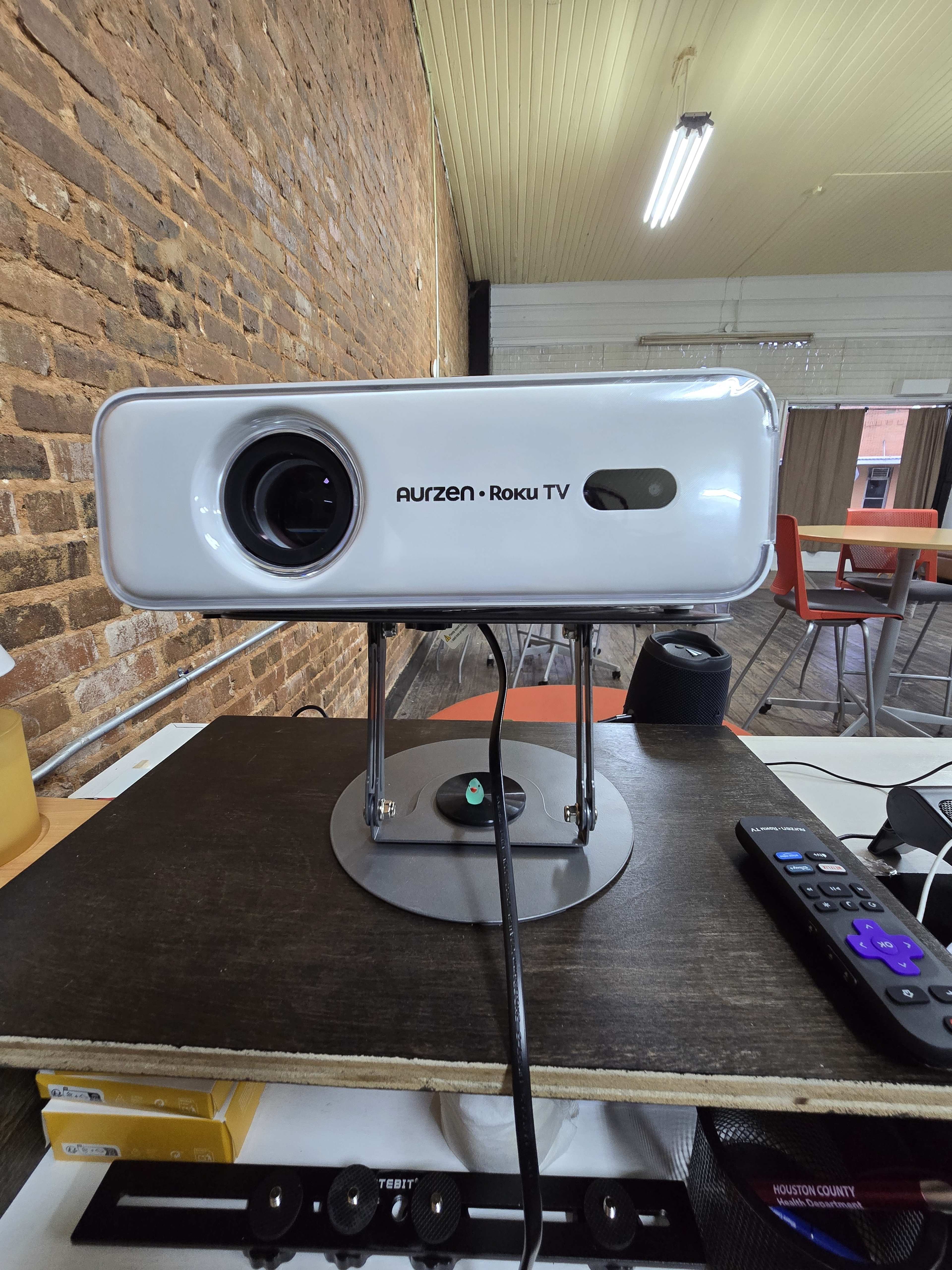 A white projector labeled "Aurzen - Roku TV" is positioned on a black table in a room with exposed brick walls and modern furniture.