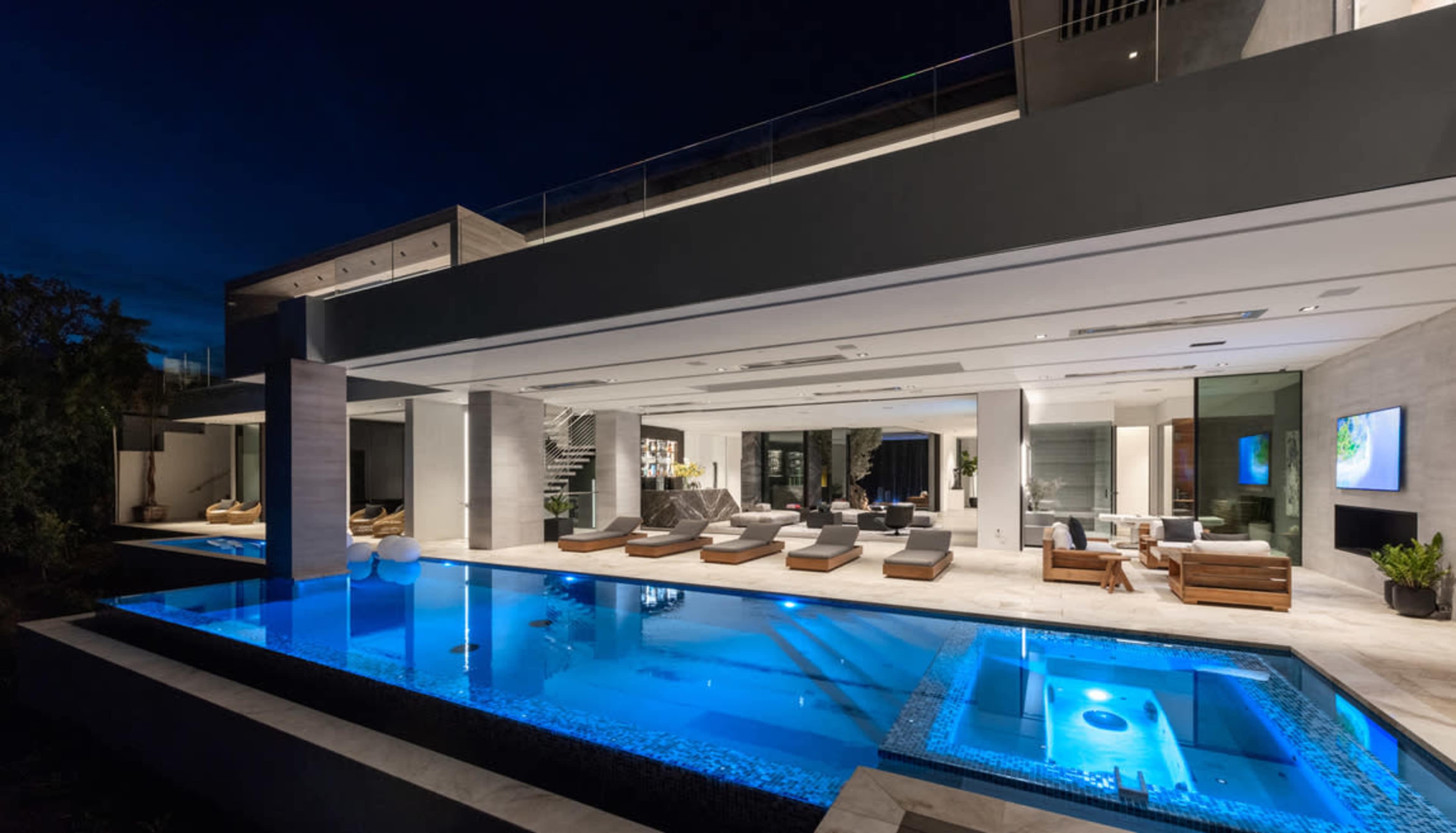 A modern house features an illuminated swimming pool with a spacious outdoor seating area and large glass walls at night.