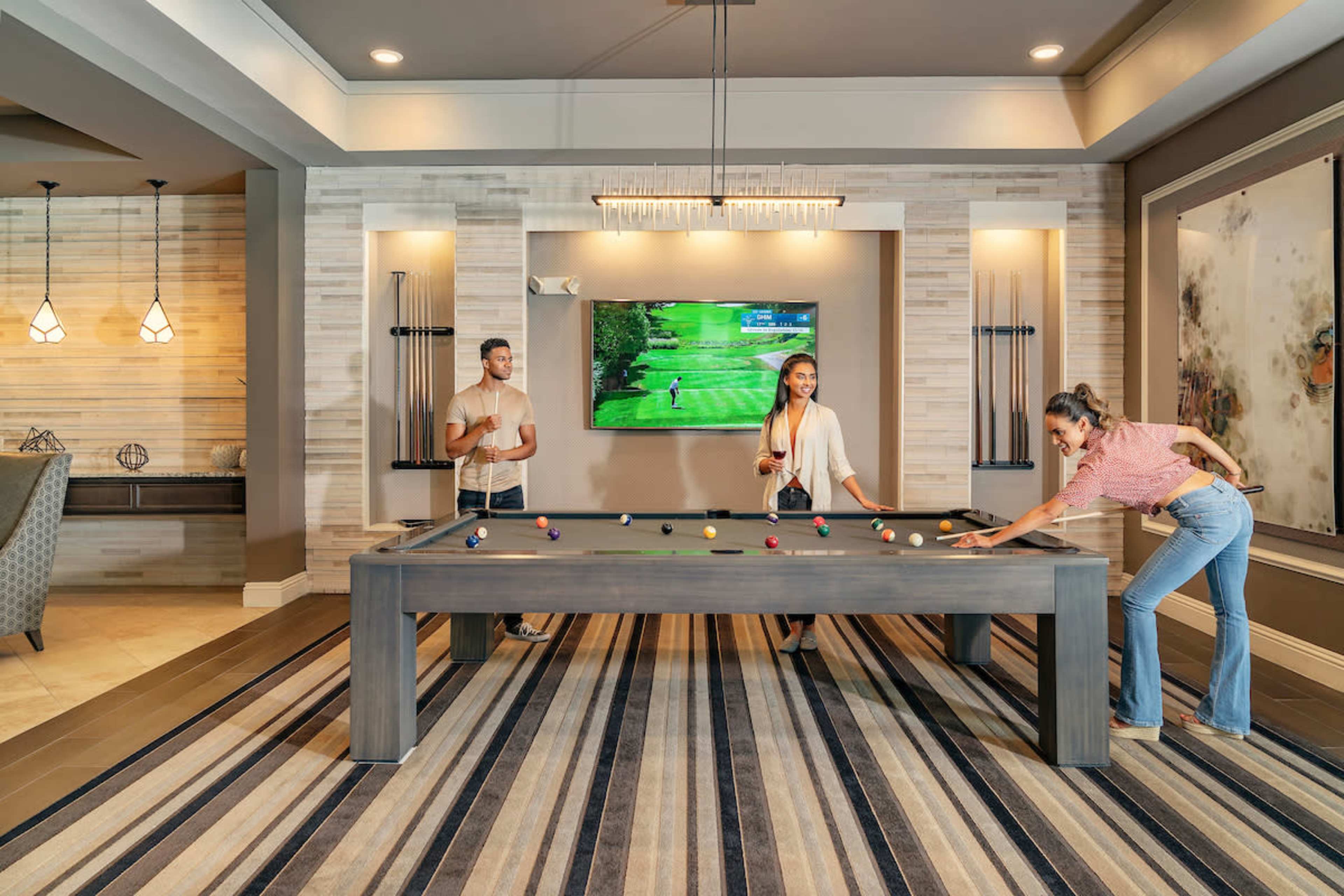 Three people are playing billiards at a modern game room with a large screen displaying a golf game in the background.