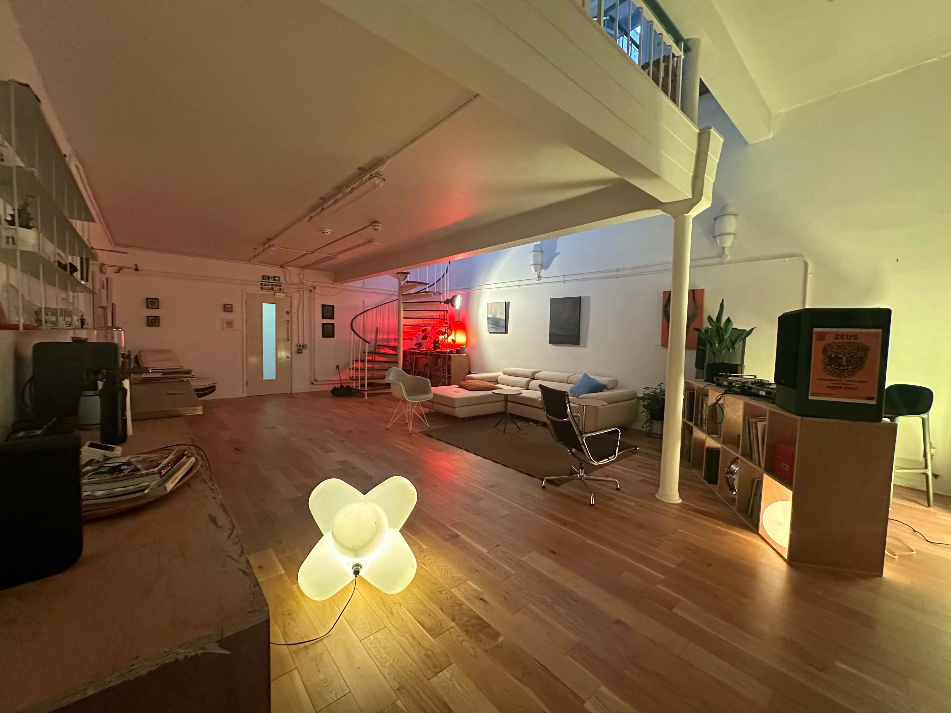 The image shows a spacious room with wooden flooring, featuring modern furniture, a spiral staircase, and a glowing light fixture in the foreground.