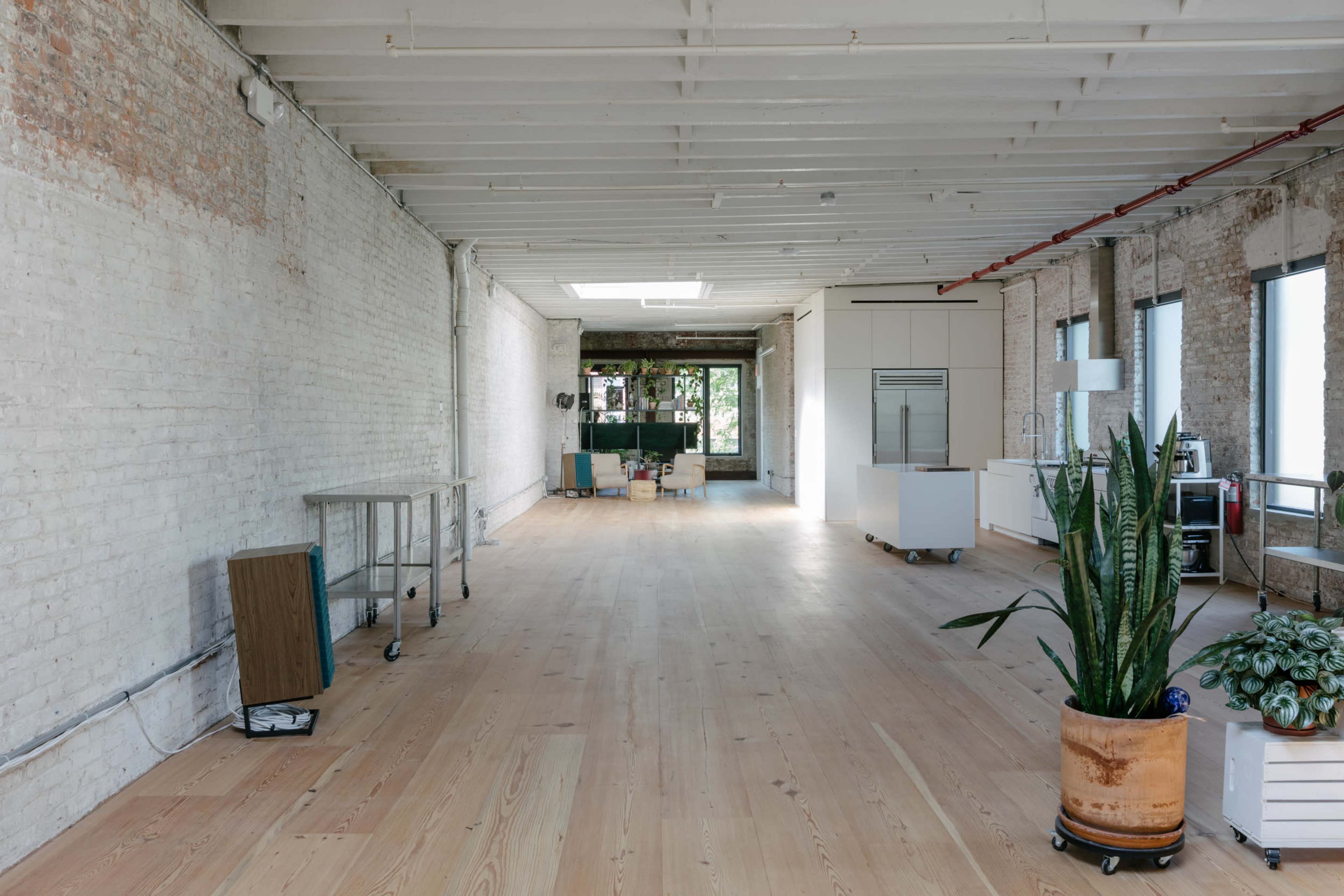 A spacious interior with wooden flooring, white brick walls, and large windows, featuring minimal furniture and a potted plant in the foreground.