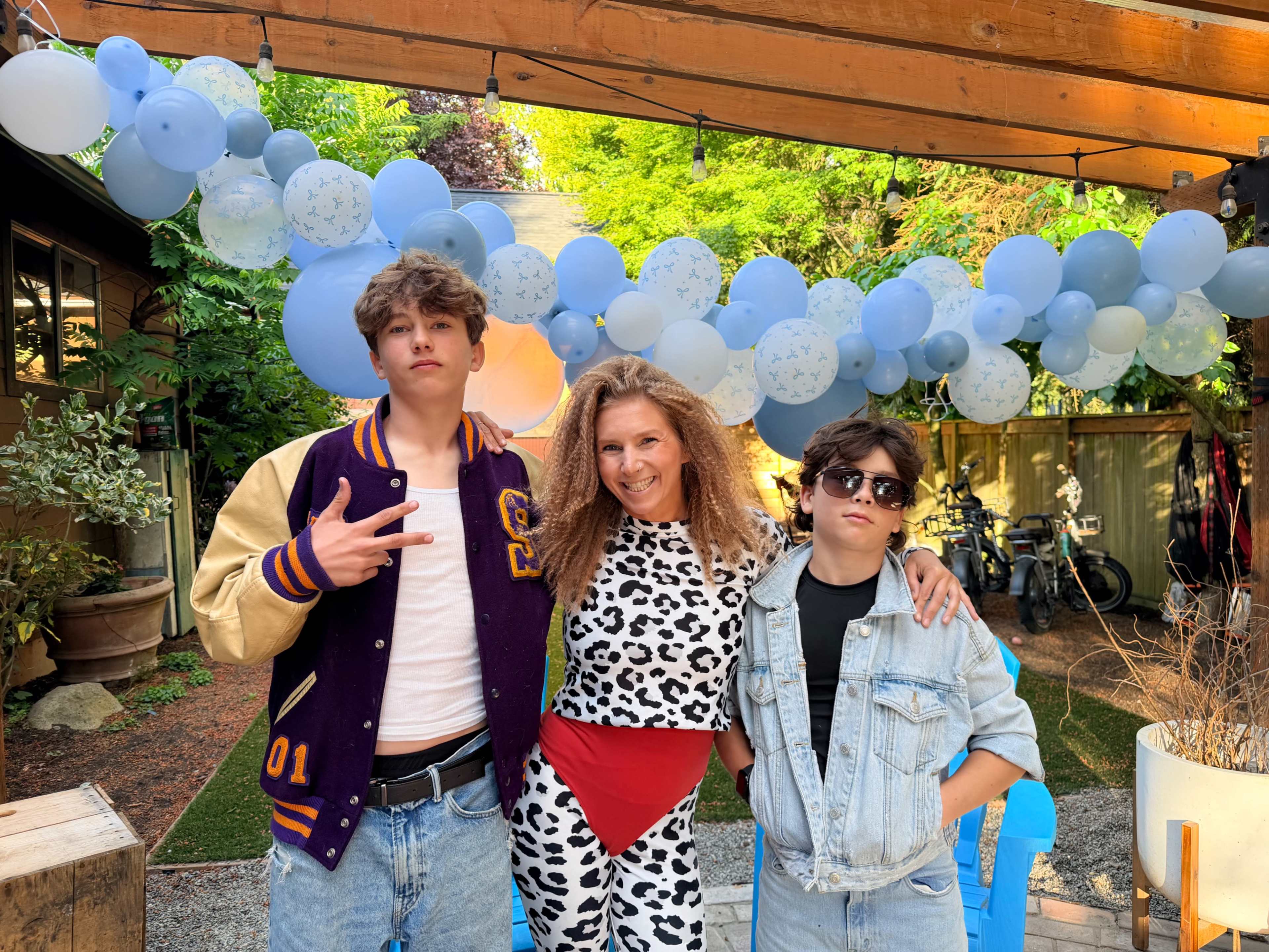 Two teenagers and an adult woman pose together under a decorative balloon arch in a backyard setting.