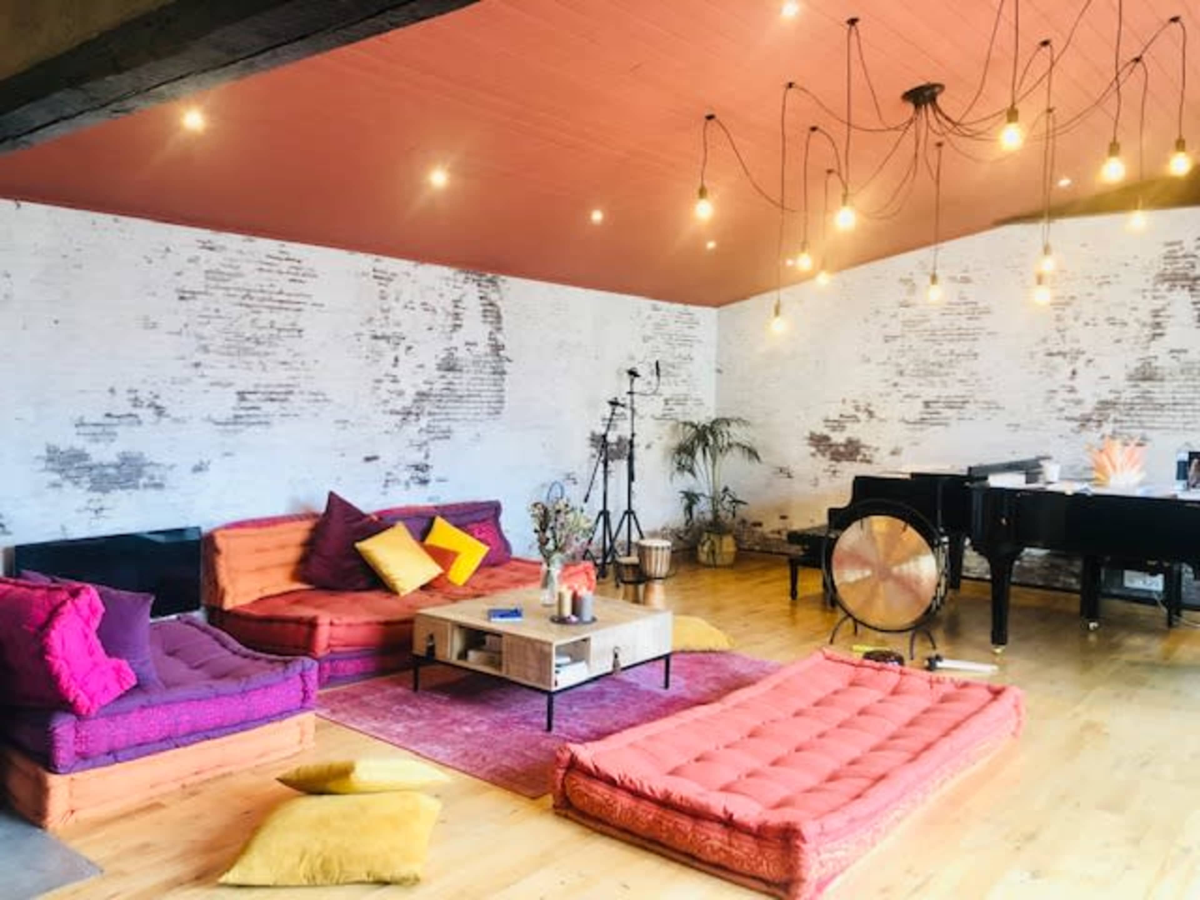 The image shows a spacious room with wooden flooring, featuring colorful floor cushions, a coffee table, a piano, and several hanging light bulbs.