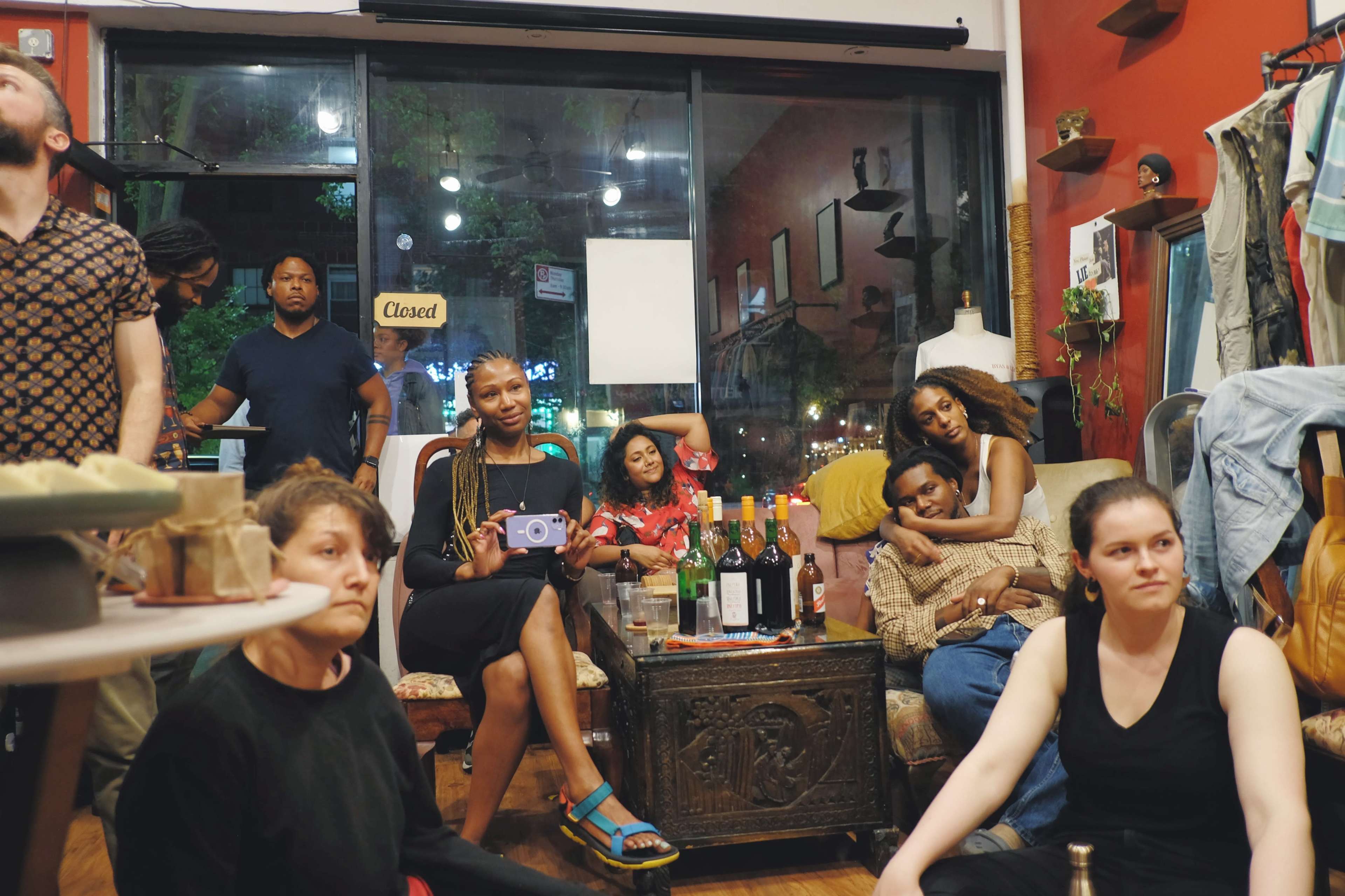 A group of people sit and stand in a warmly lit room filled with bottles of wine and clothing displays, engaged in conversation and relaxation.