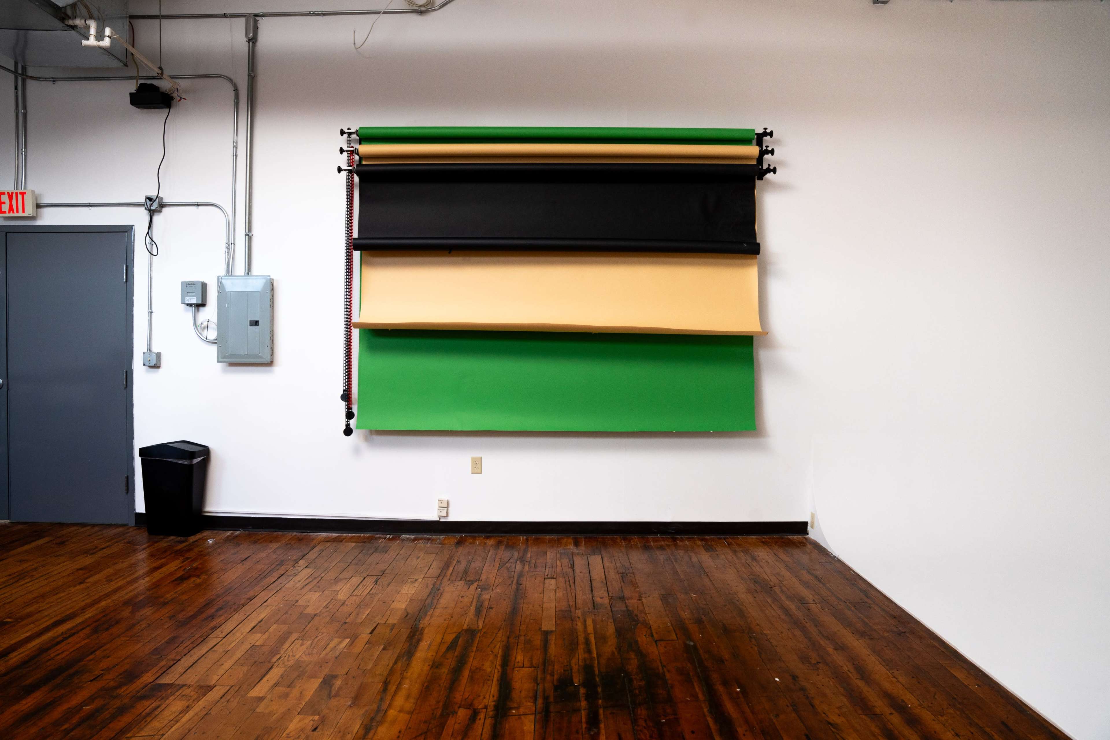 A wall with four rolls of backdrop paper in various colors arranged above a wooden floor in a spacious room.