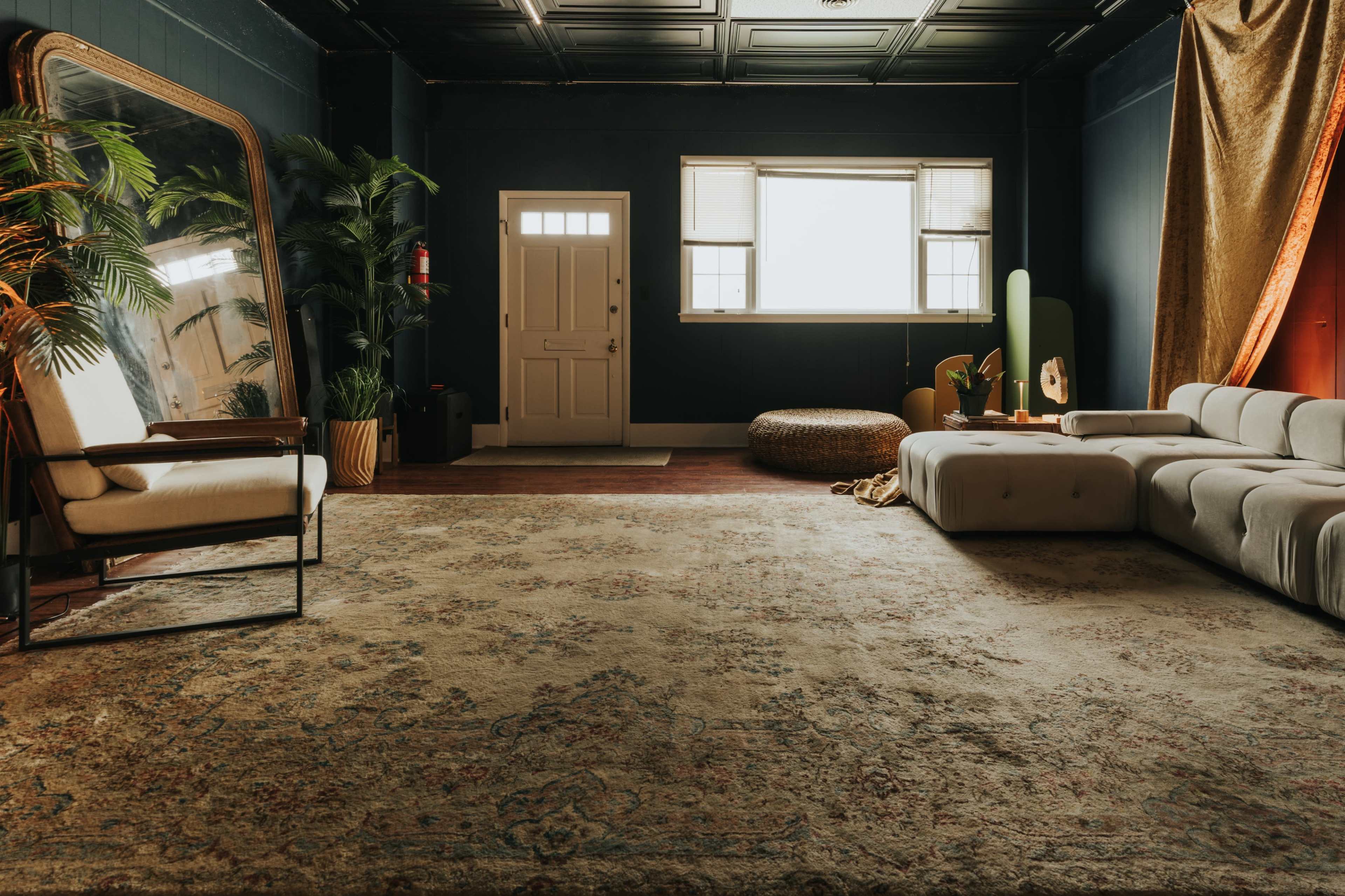 A spacious room with a large rug, a beige sectional sofa, a reflective mirror, potted plants, and a door leading outside.