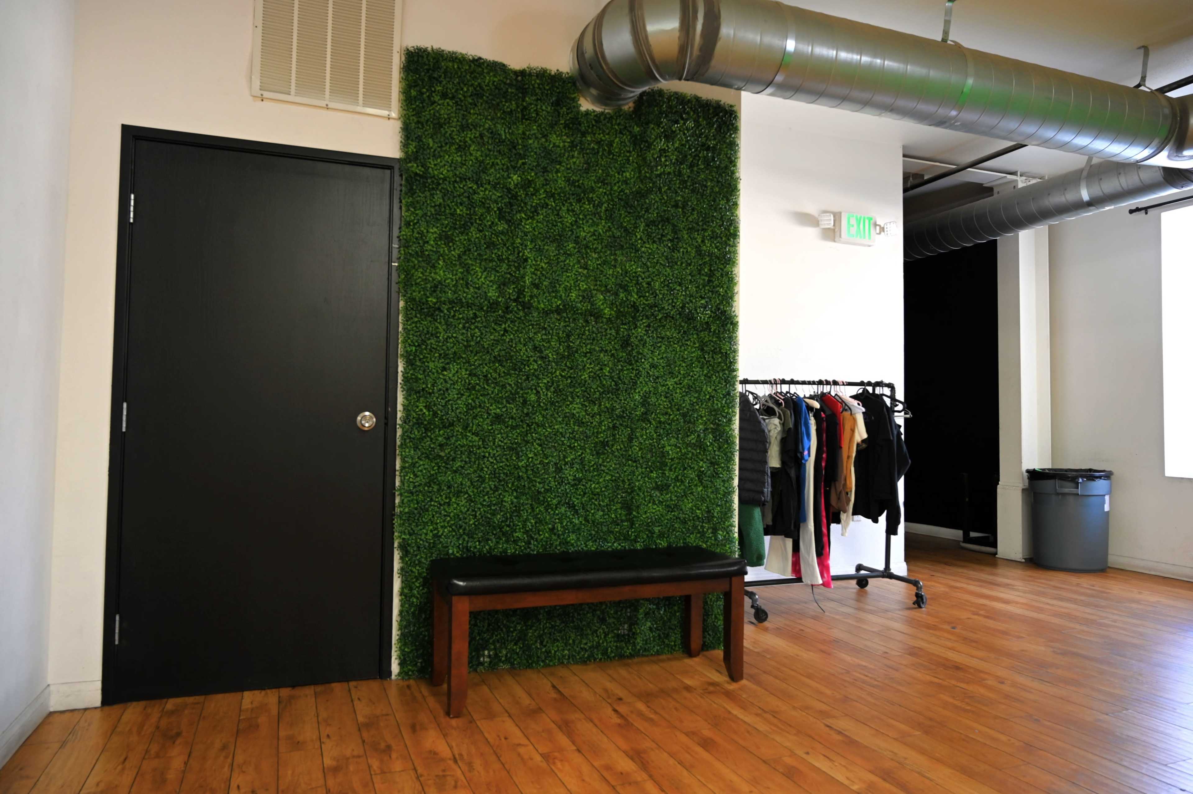 A modern interior space with a green artificial grass wall, a black door, a wooden bench, and a clothing rack filled with assorted garments.