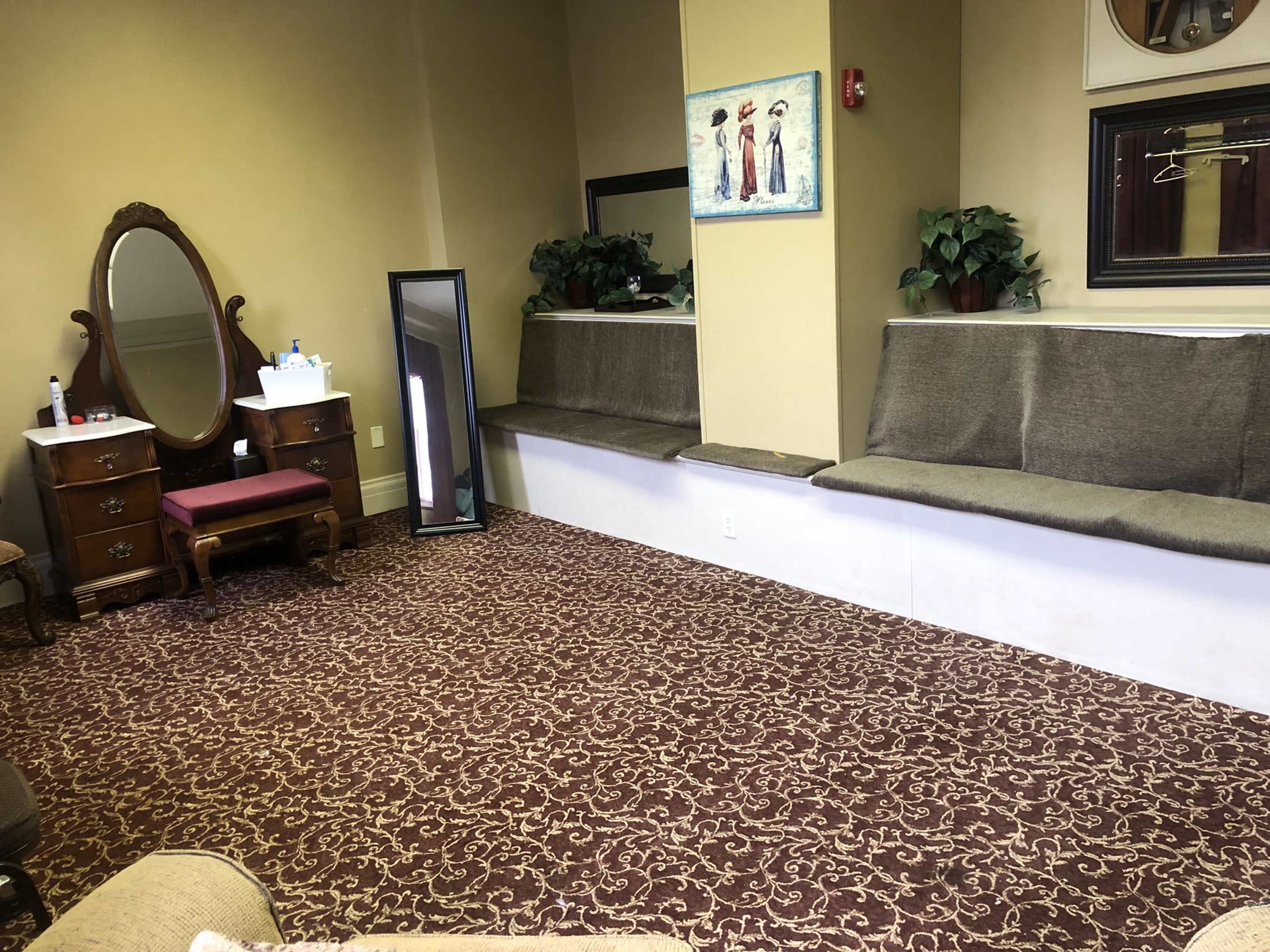A spacious room with a patterned carpet, a vintage dresser, a mirror, and two seating areas along the wall.