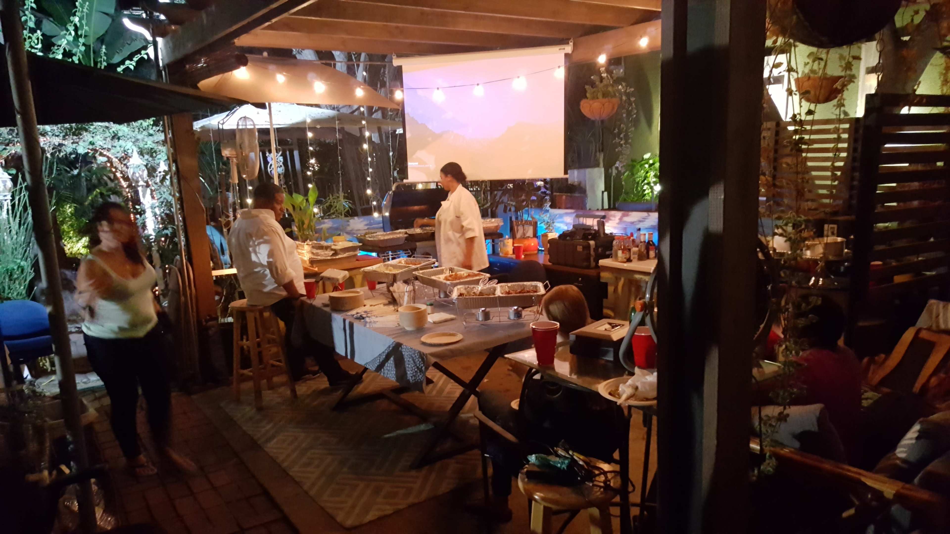 A backyard gathering features people preparing food at a table under string lights and a projected screen.